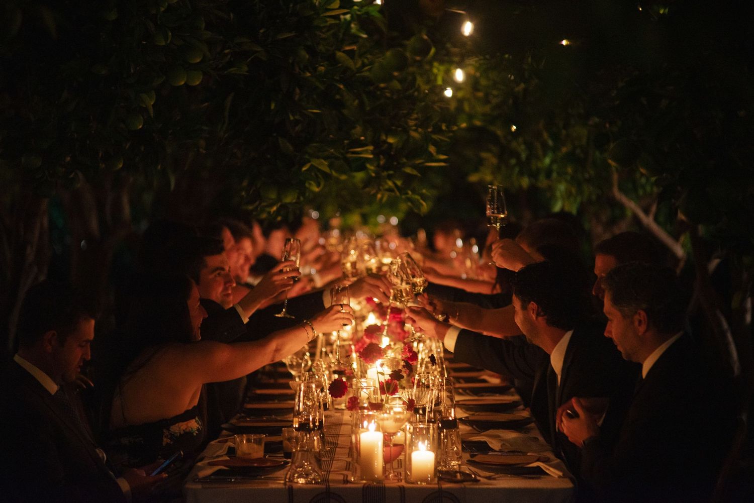 Guests raise glasses in a toast across a long candlelit outdoor dinner table decorated with flowers under trees at night.