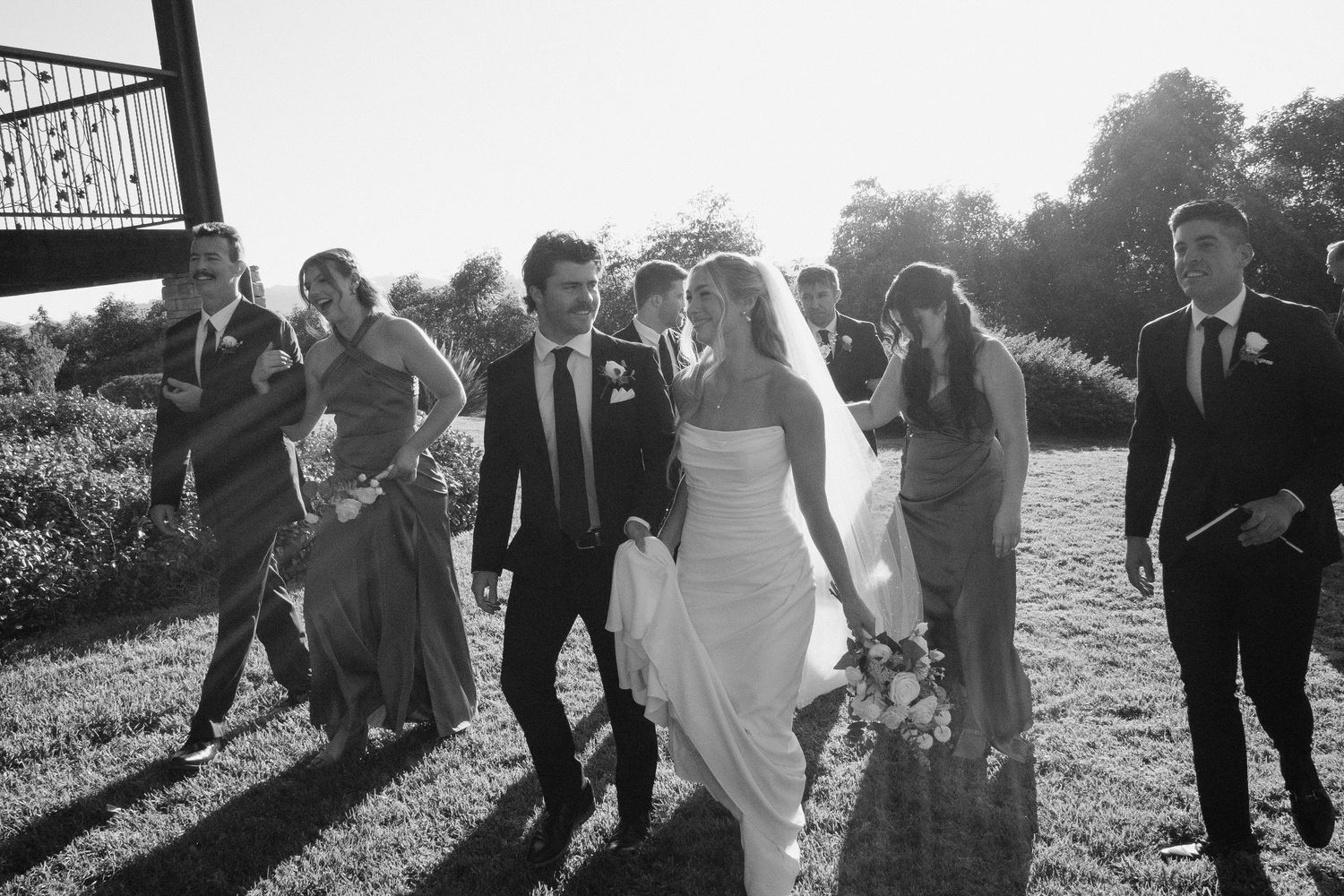 Black and white photo of a bride and groom walking with their wedding party outdoors on a sunny day.