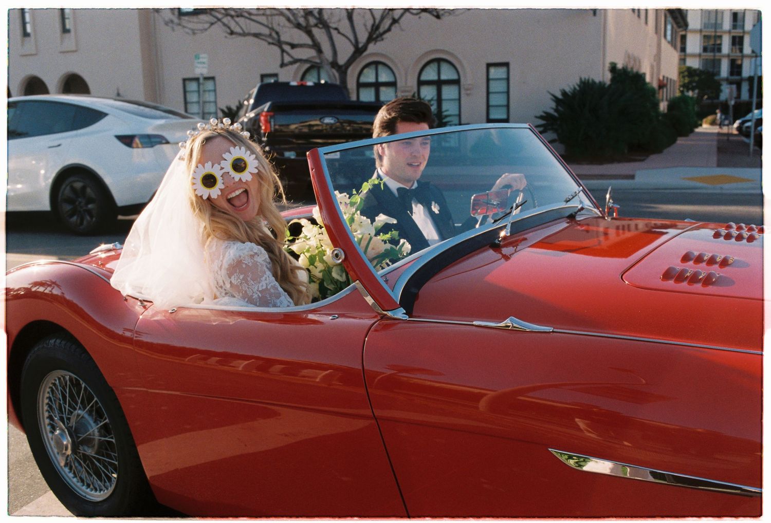 Bride wearing floral crown and groom in suit ride in a vintage red convertible sports car on wedding day.