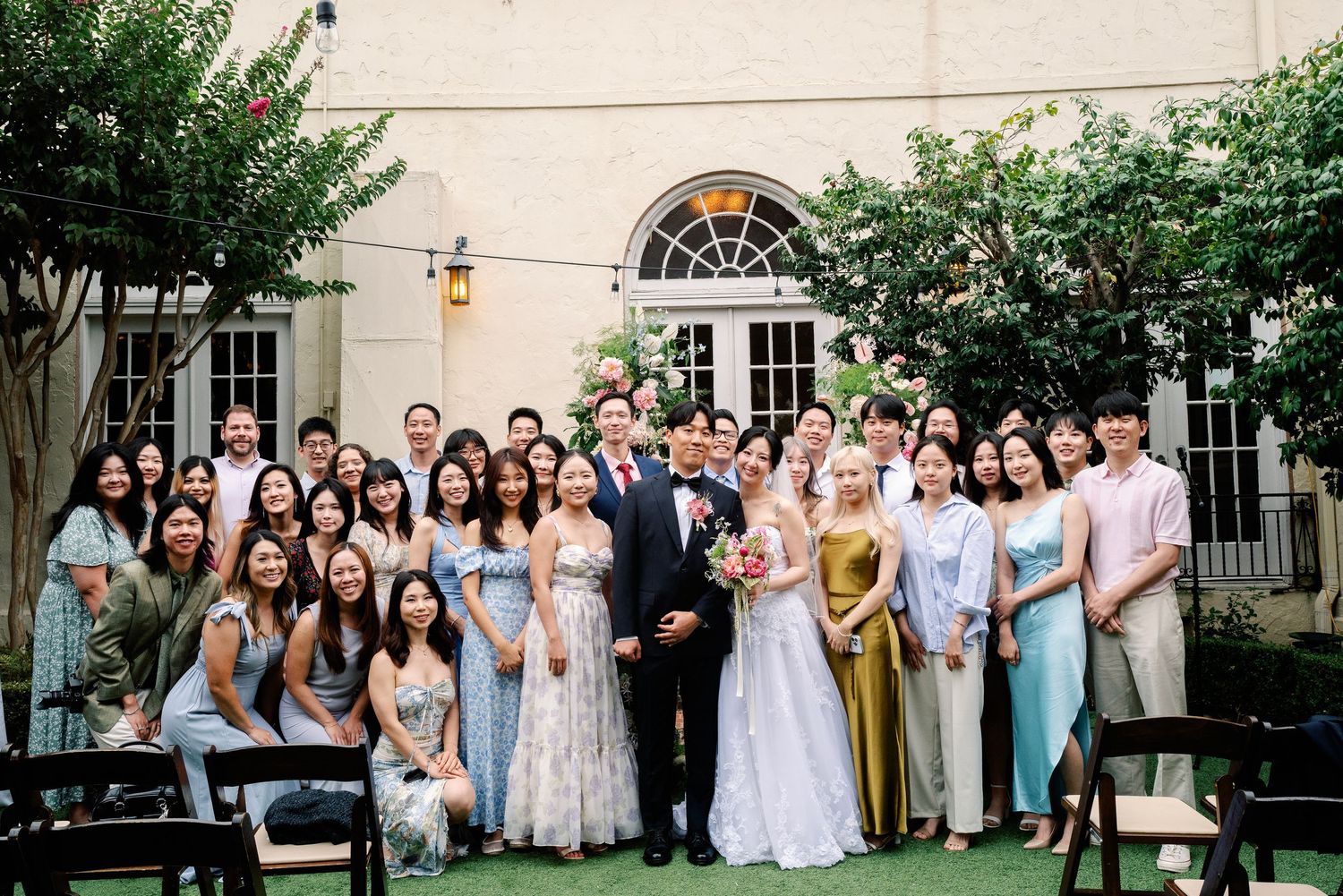 Wedding couple poses with large group of guests in sunny outdoor courtyard ceremony venue.