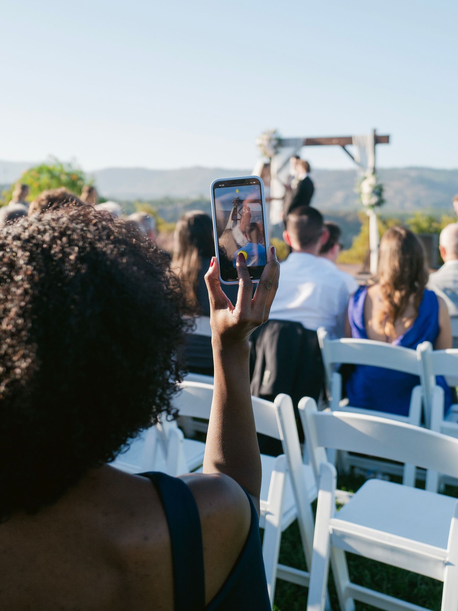 Wedding guest capturing bride and groom at altar on smartphone during sunny outdoor ceremony.