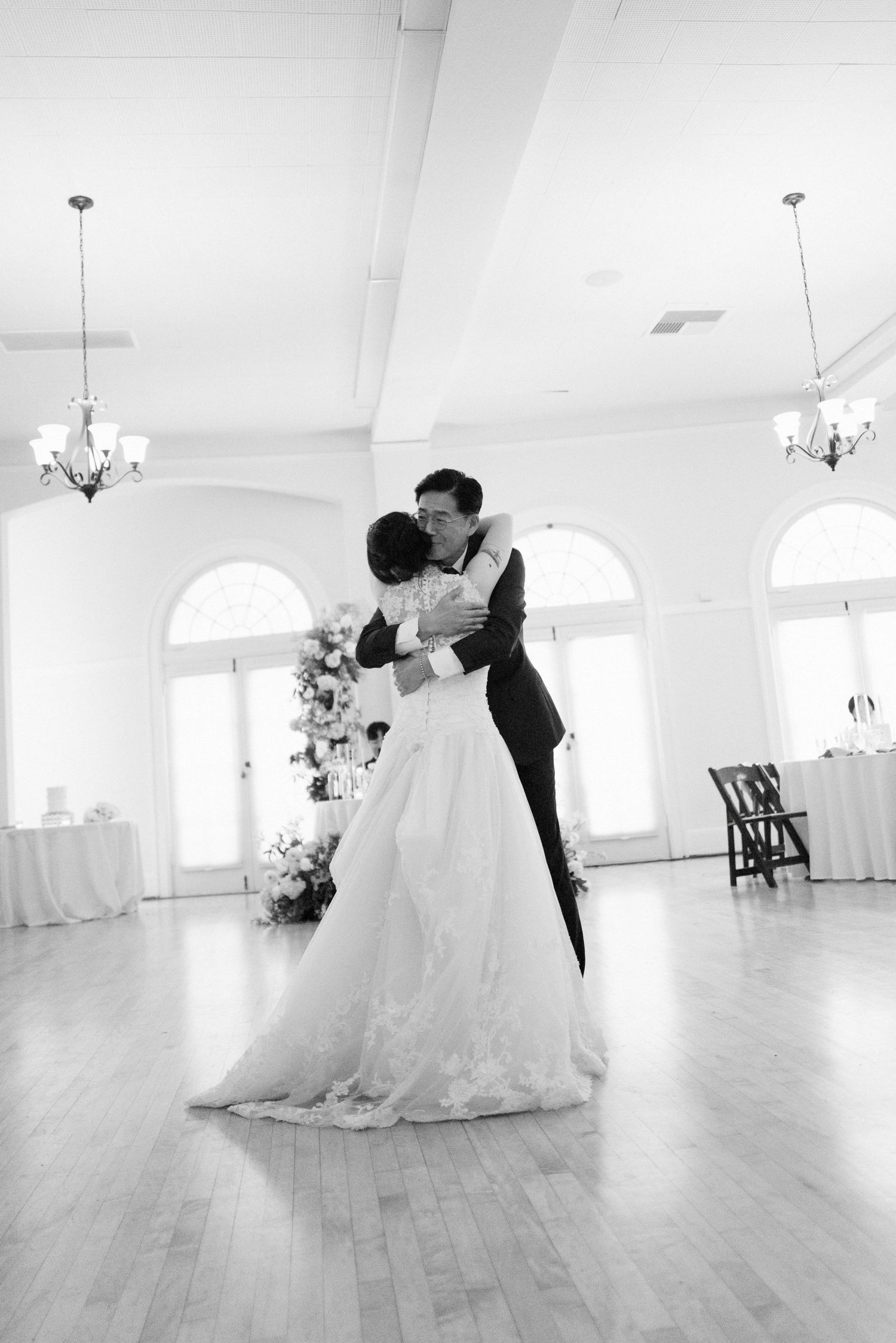Black and white photo of bride and groom sharing first dance in elegant ballroom with chandeliers and arched windows.