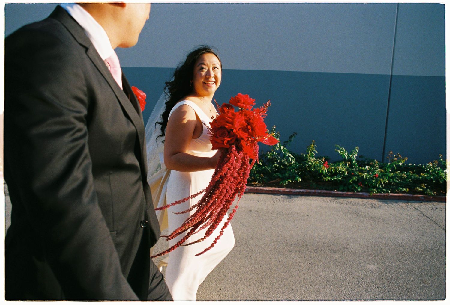 Bride in white dress holding red bouquet smiles at groom in black suit outdoors on wedding day.