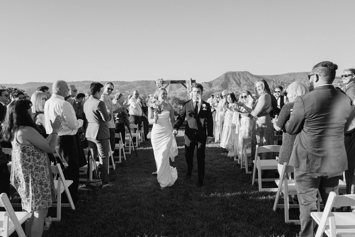 Newlyweds walking down aisle as guests celebrate at scenic outdoor wedding ceremony in black and white.