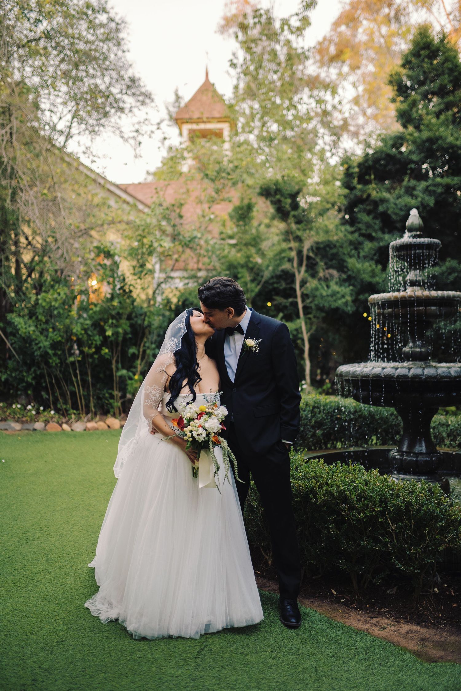 Bride and groom share a romantic kiss near a stone fountain in a lush garden wedding venue.