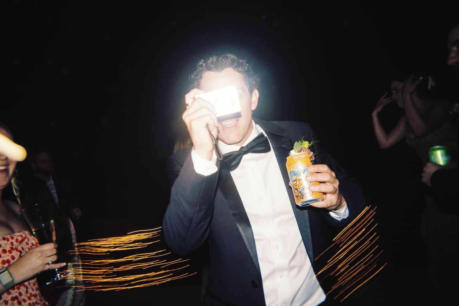 Man in tuxedo takes photo with flash while holding a drink at dark outdoor wedding reception.