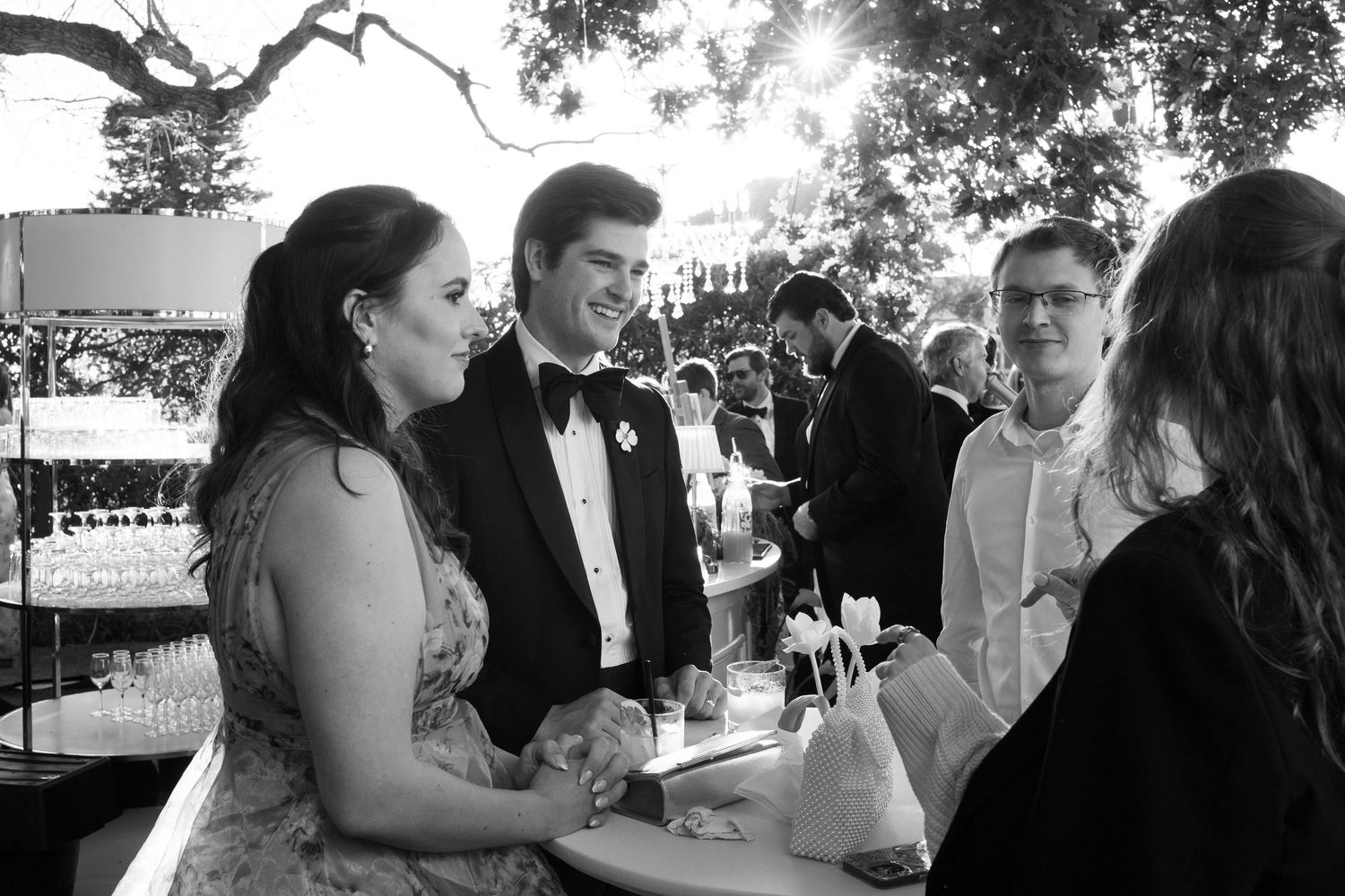 Elegantly dressed wedding guests smile and converse at an outdoor cocktail reception in black and white.