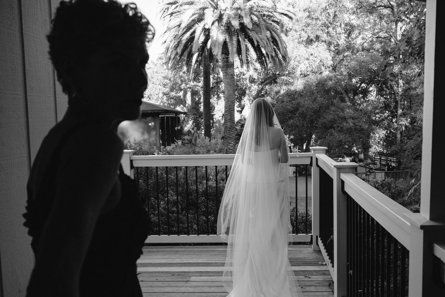 Black and white photo of a bride in a long veil standing on a wooden deck, facing palm trees, as a silhouetted person watches from the left.