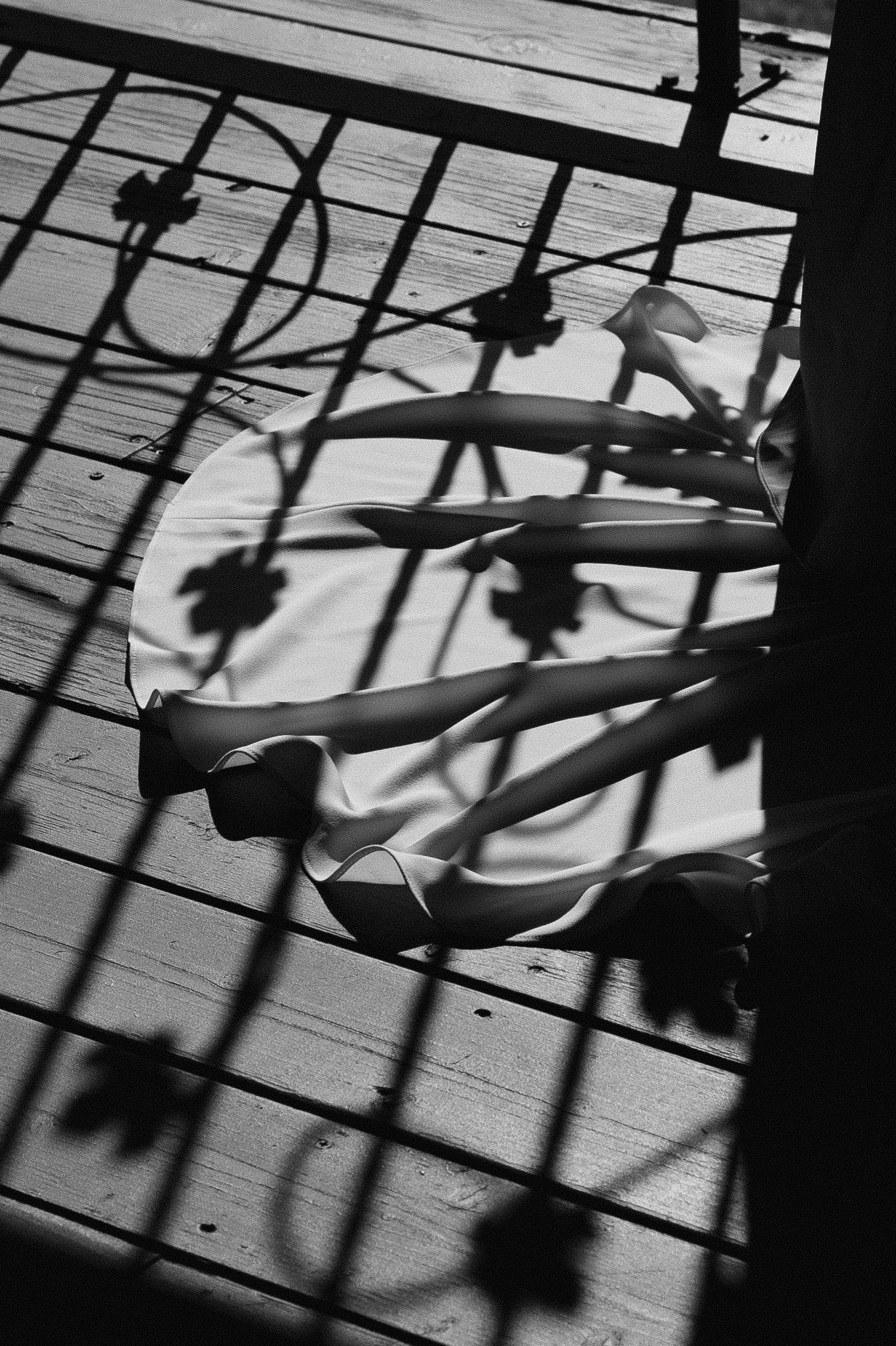 Black and white photo of a bride's legs and wedding dress hem casting shadows on a wooden deck through ornate iron railings.