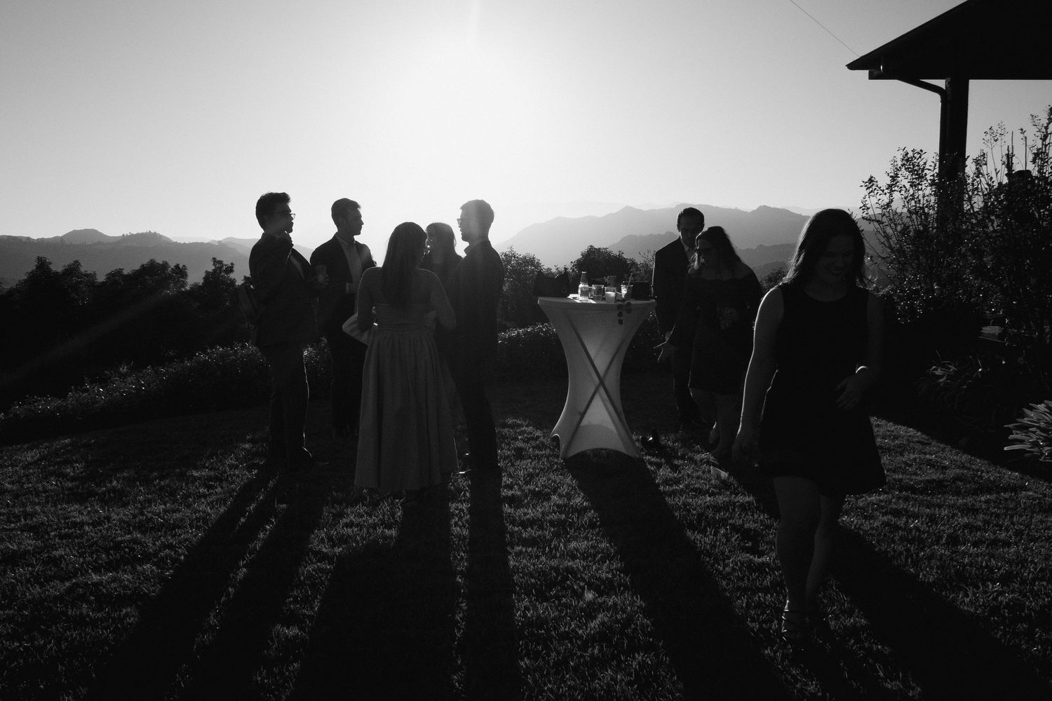 Silhouetted wedding guests standing in a sunlit garden with mountain backdrop at golden hour in black and white.