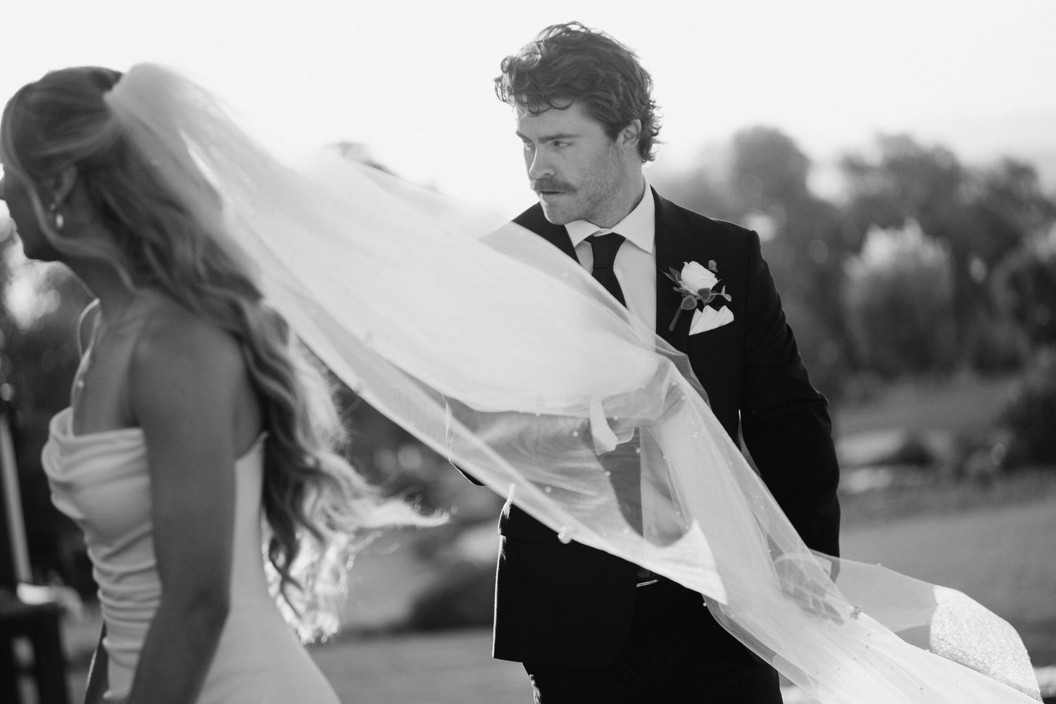 Black and white photo of groom catching brides flowing veil in the wind during outdoor wedding.