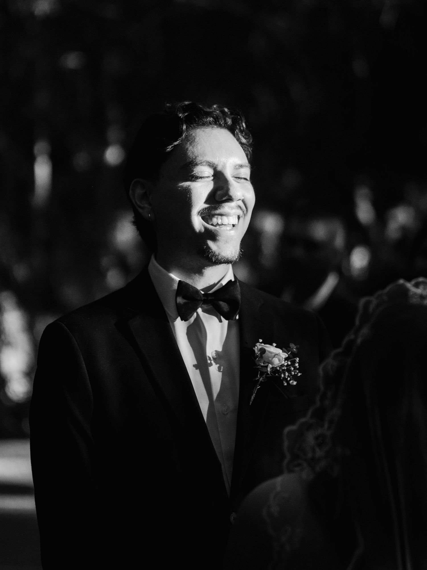 Groom in black tuxedo and bow tie smiling joyfully during outdoor wedding ceremony, black and white photo.