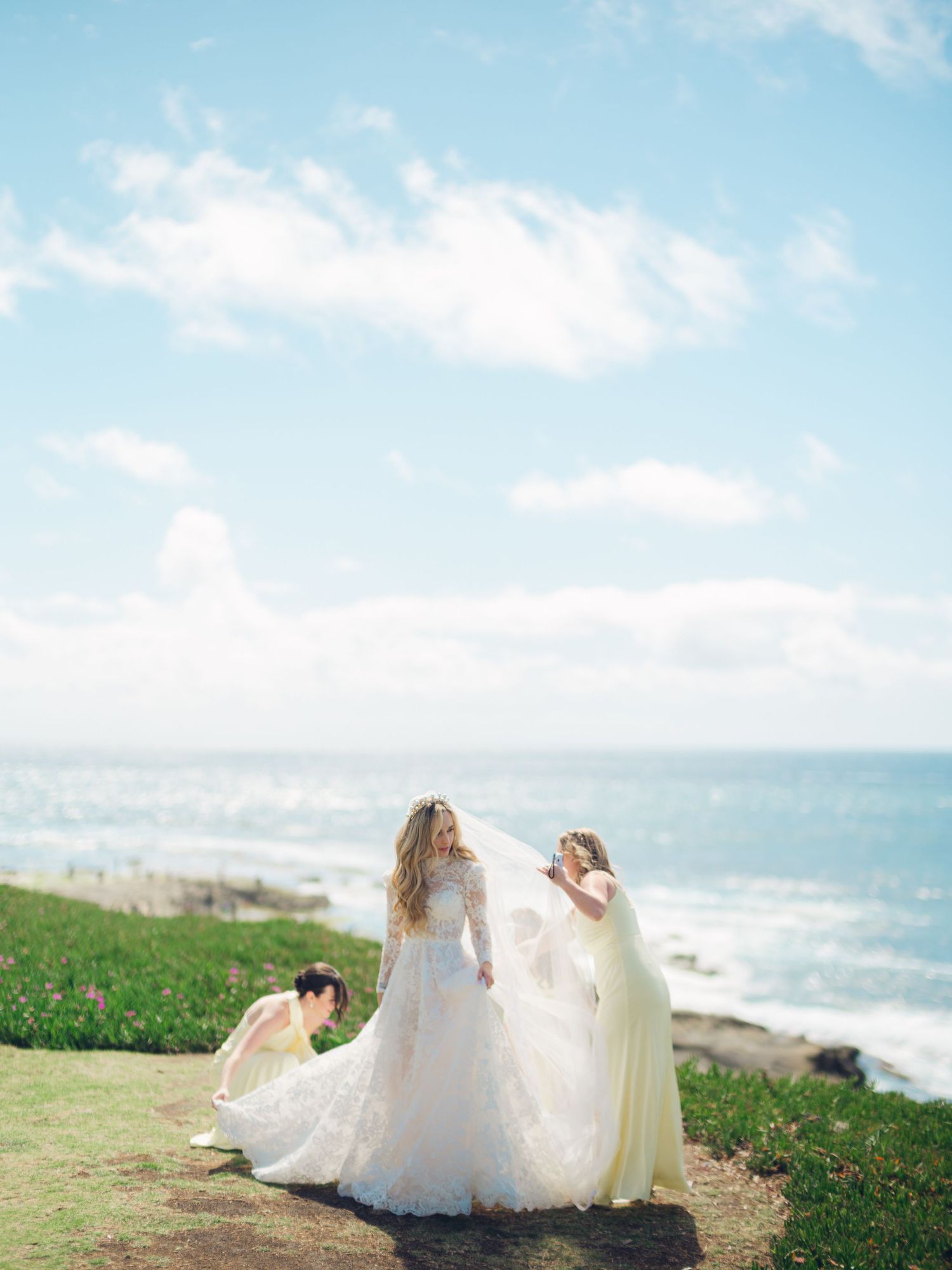 Bridesmaids arrange bride's flowing veil on a coastal cliff with the ocean in the background.