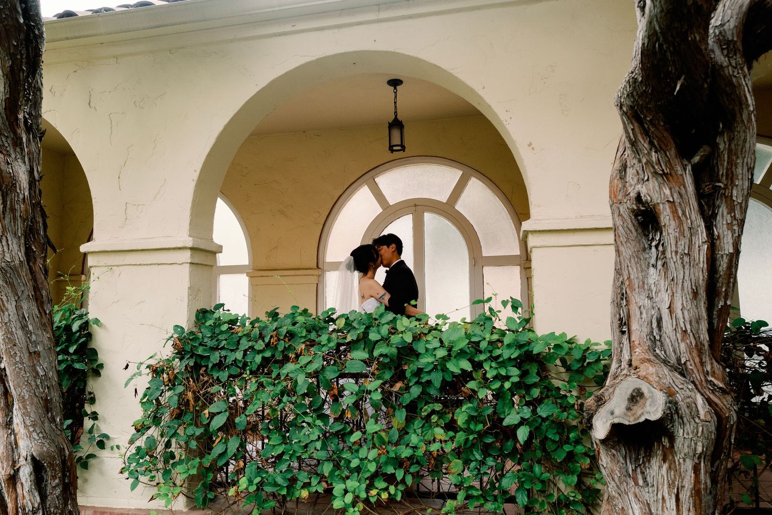 Bride and groom sharing romantic kiss on ivy-covered balcony with arches.