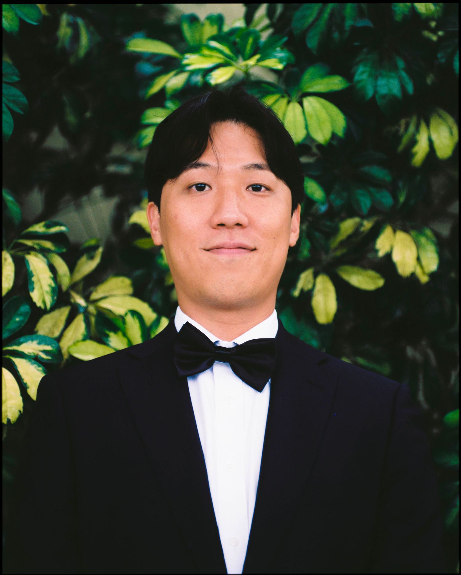 Groom in black tuxedo and bow tie smiling in front of green foliage.