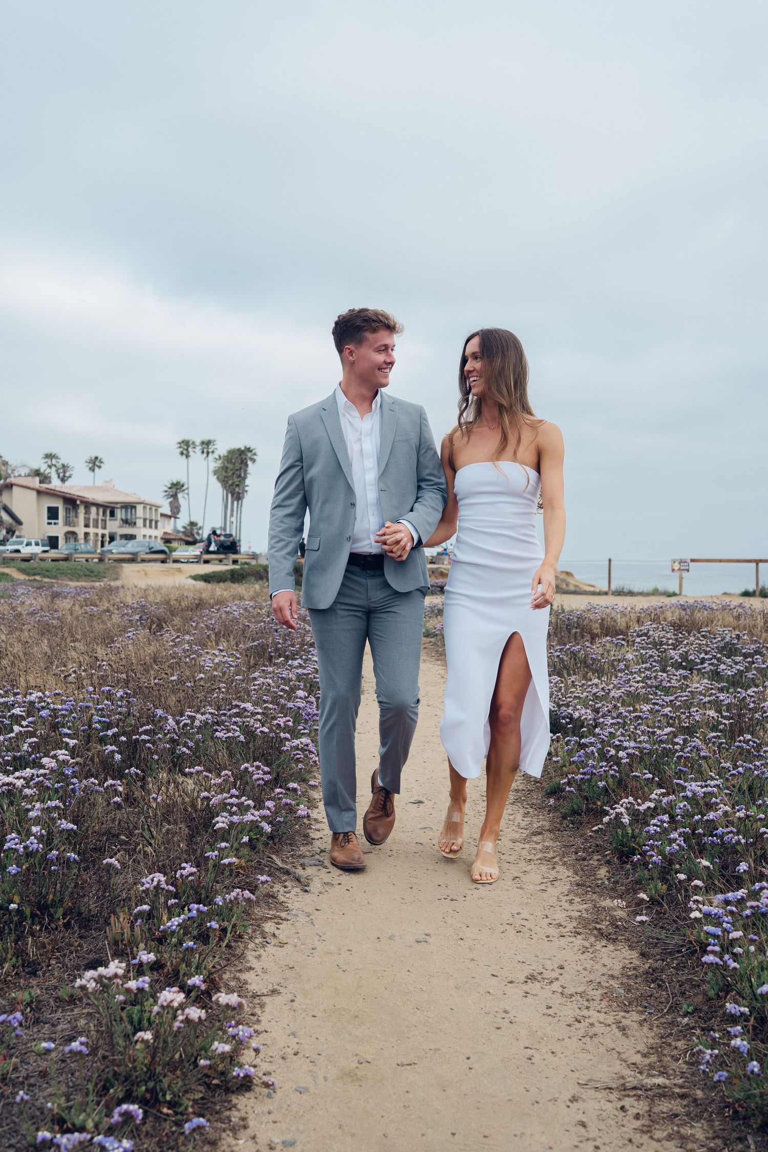 Engaged couple holding hands walking on coastal path surrounded by purple wildflowers, woman in white dress and man in gray suit.