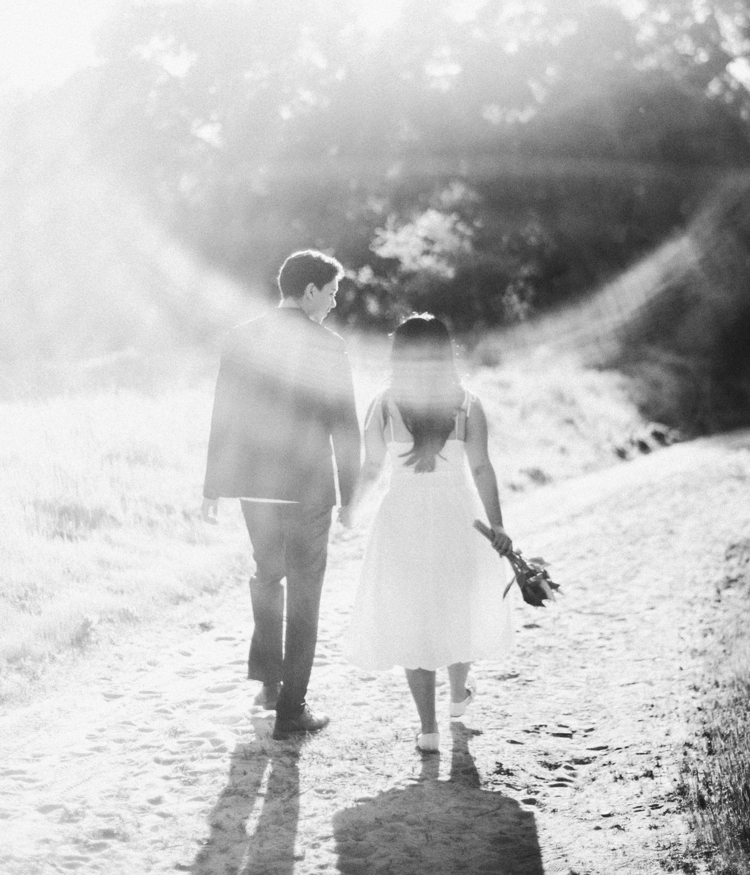 Bride and groom walk hand in hand down sunlit path in dreamy black and white photo.