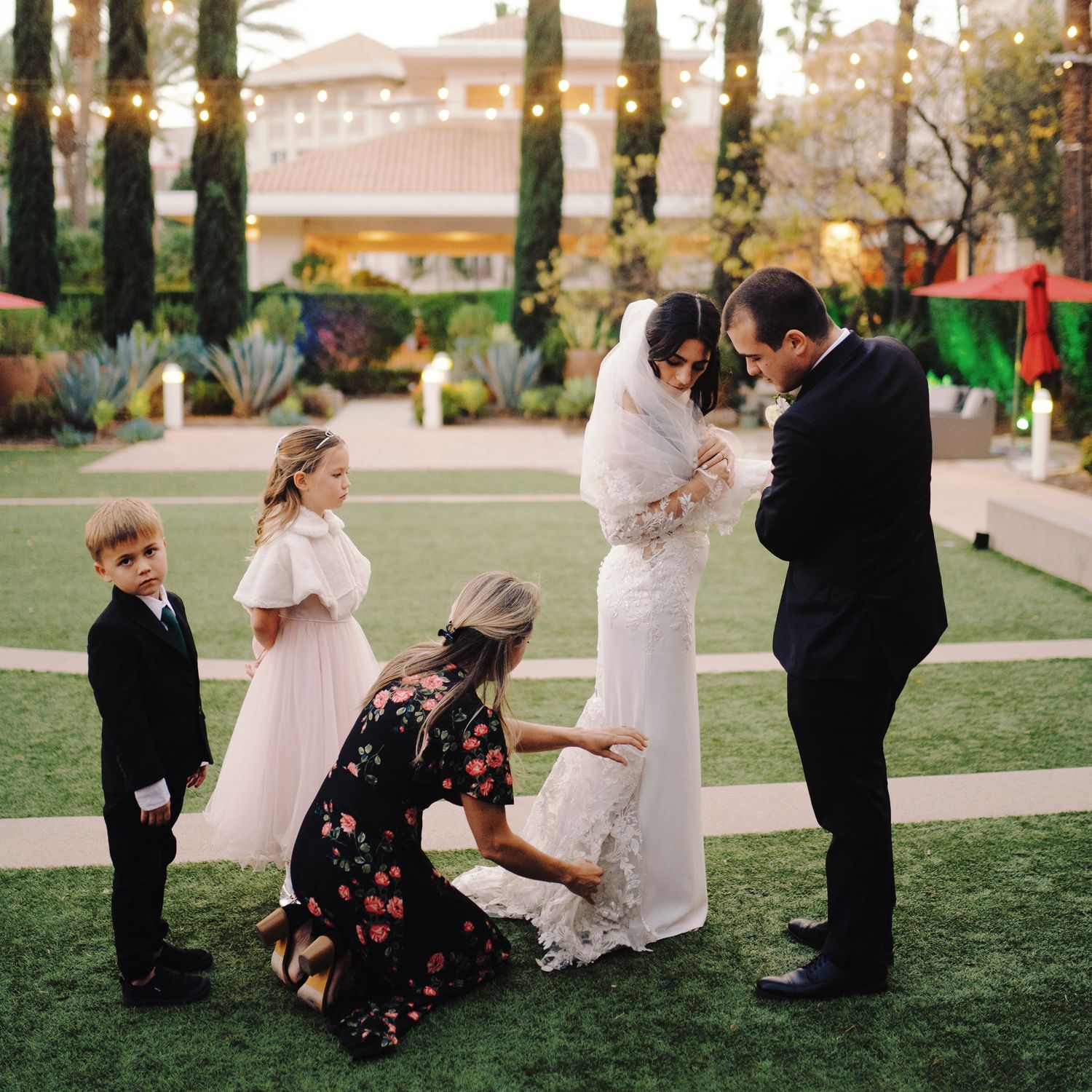 Bride in white gown with veil being adjusted by wedding party outdoors at elegant venue with string lights.