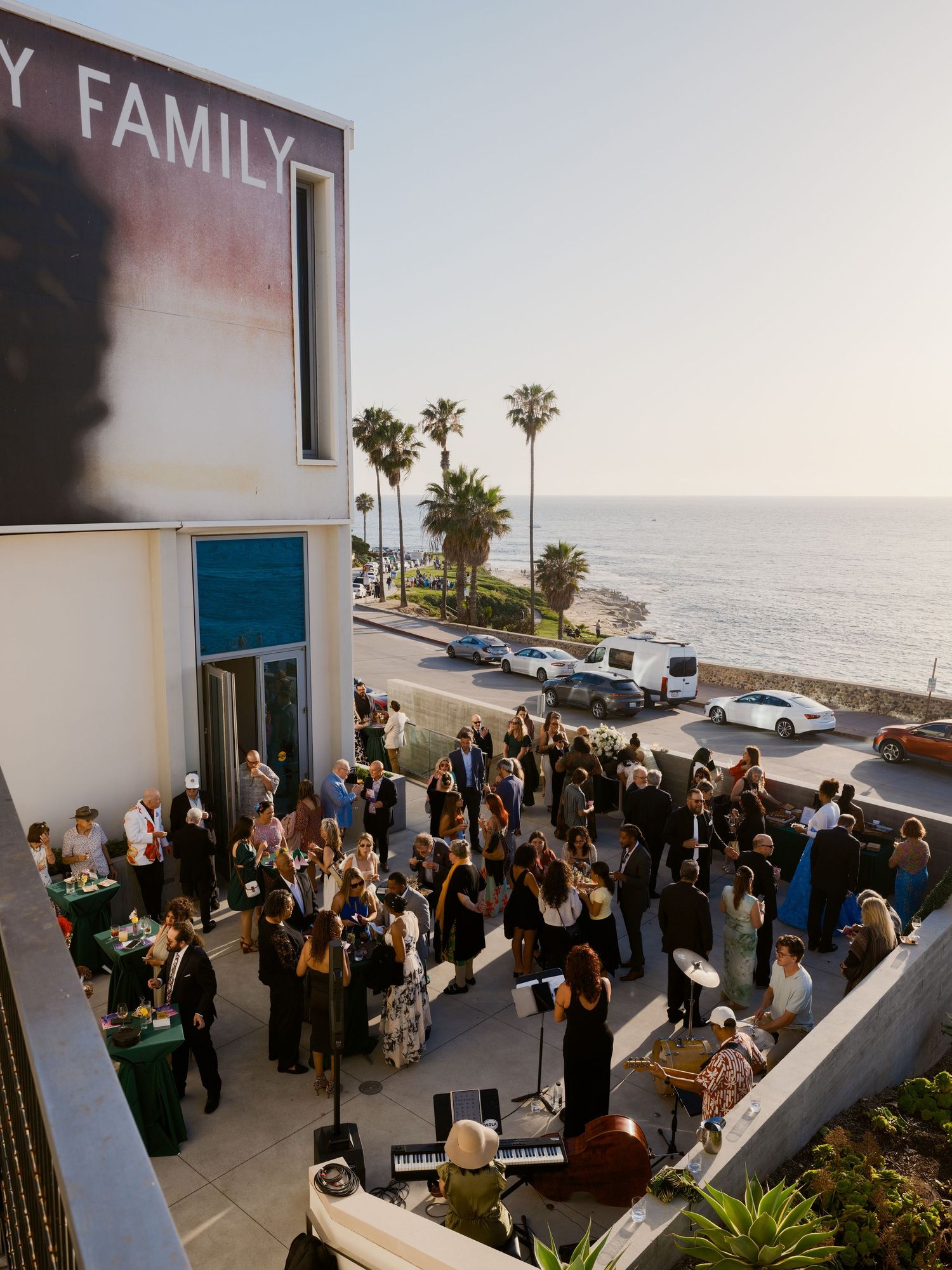 Outdoor coastal event with guests mingling on a terrace, palm trees, and ocean views under a building sign.
