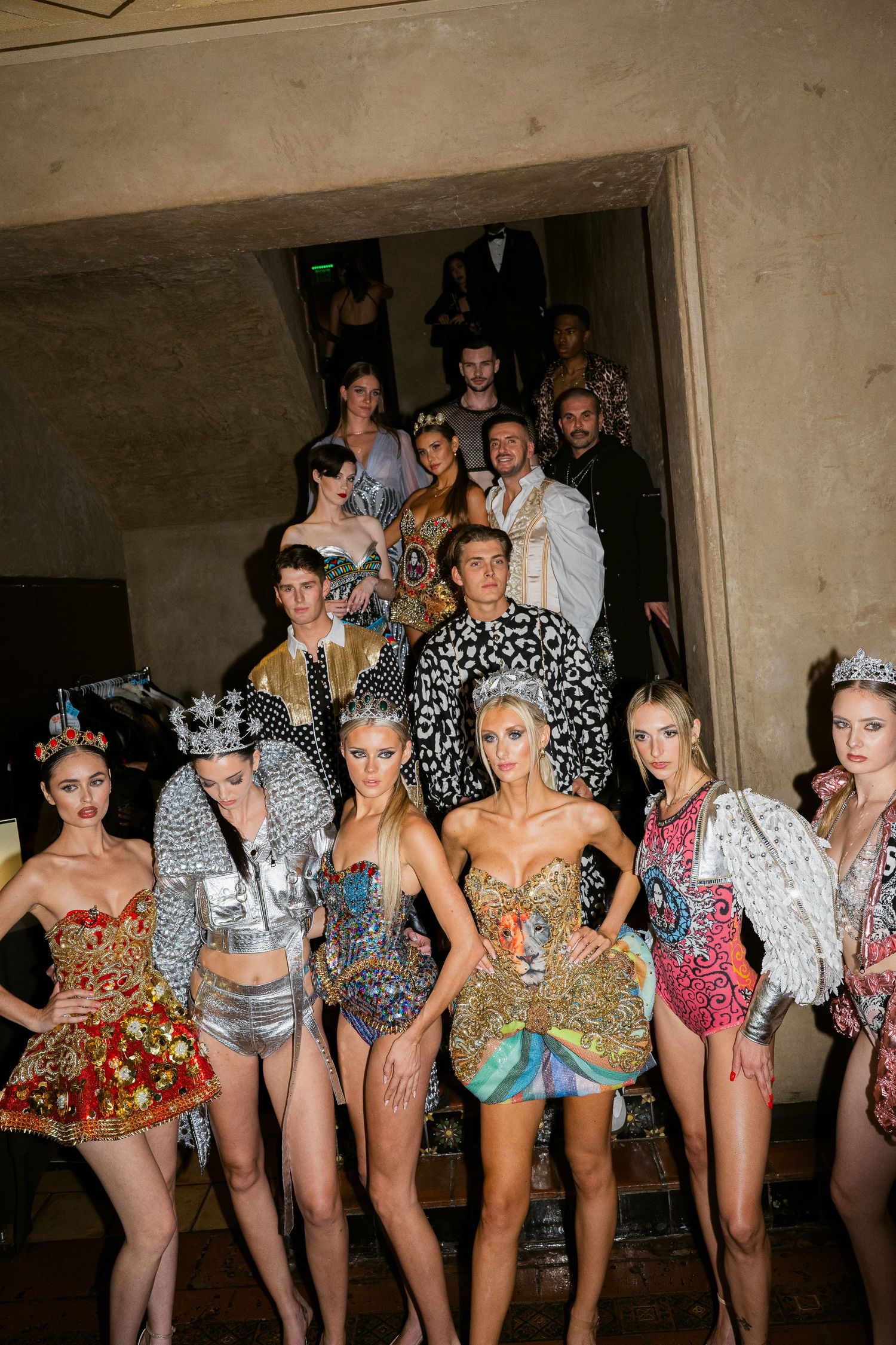 Models pose in elaborate, bejeweled fashion ensembles on a staircase at a glamorous runway event.