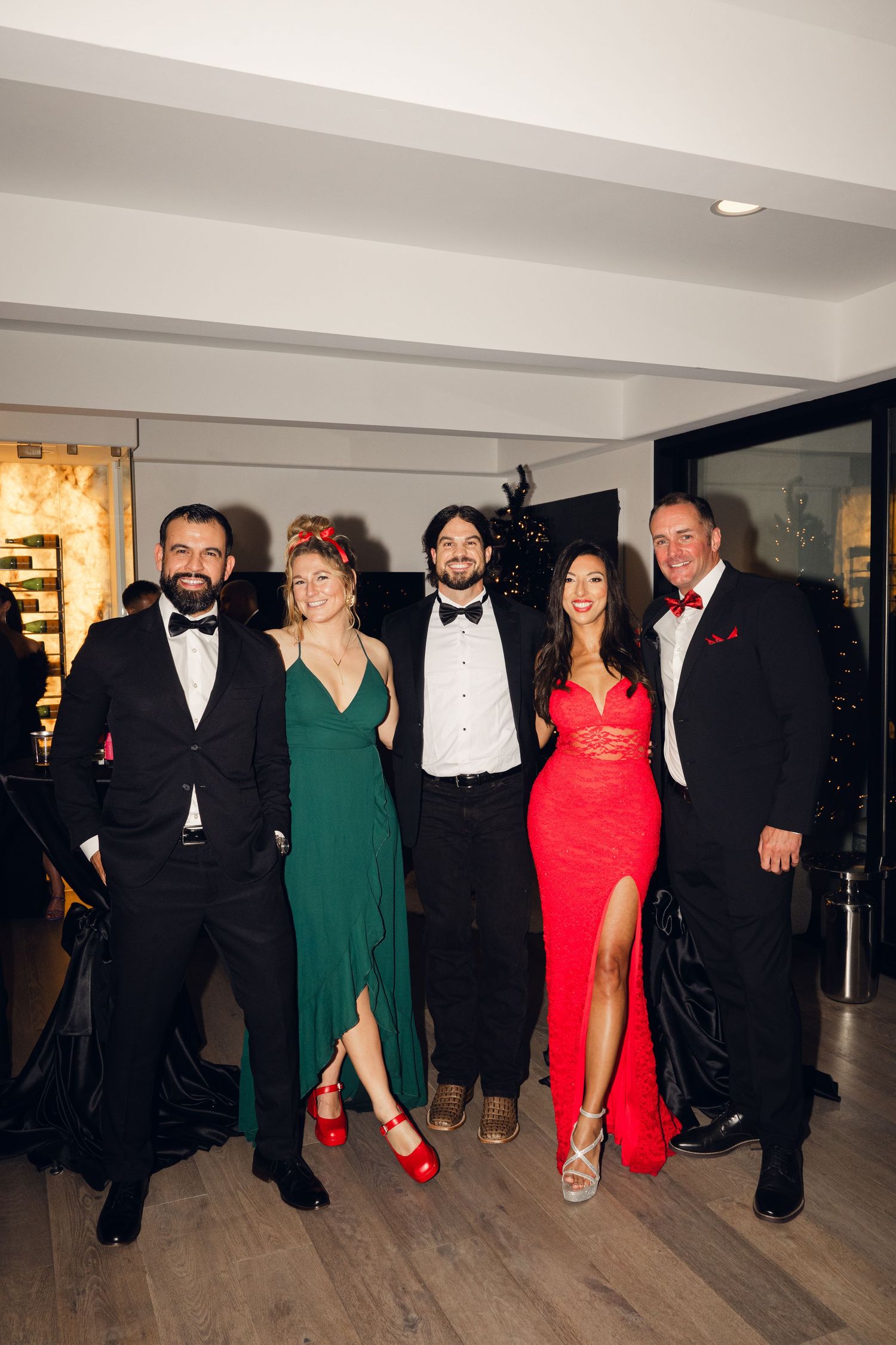 Five elegantly dressed people posing at a formal event, wearing black tuxedos and colorful gowns in green and red.