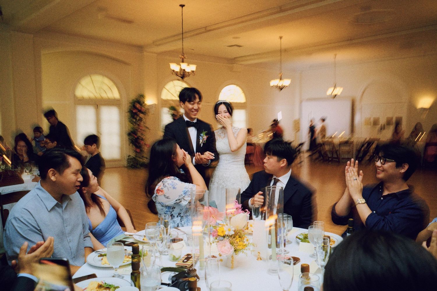 Bride and groom greeting guests at elegant wedding reception dinner table with candlelight and chandelier.