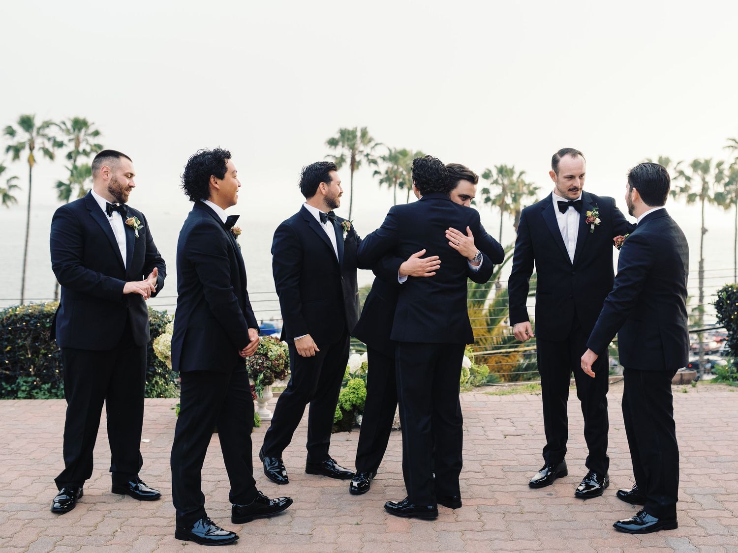Groomsmen in black tuxedos sharing embraces outdoors with palm trees and ocean view at a wedding.