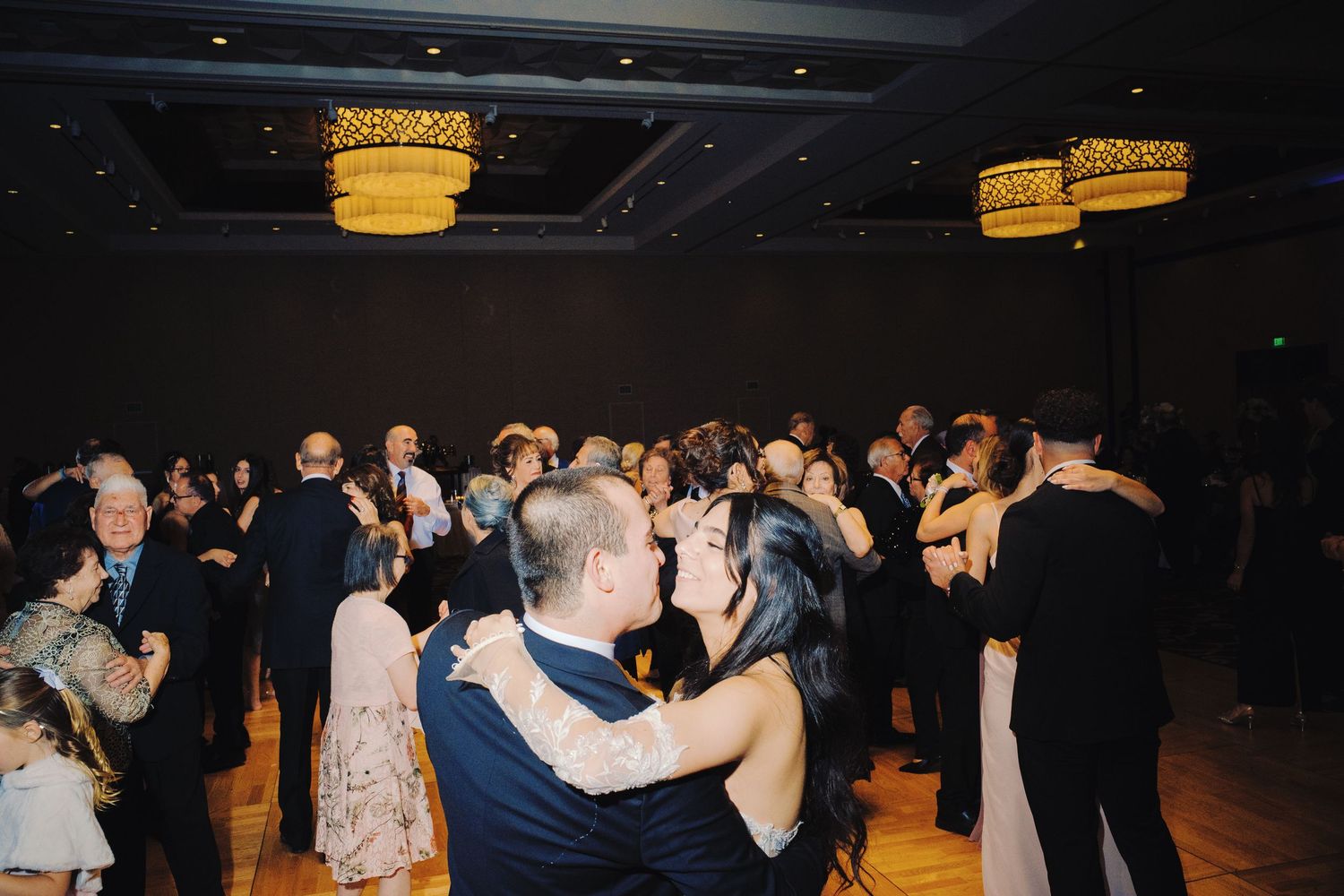 Couples dancing together at an elegant wedding reception with warm golden chandeliers illuminating the ballroom dance floor.