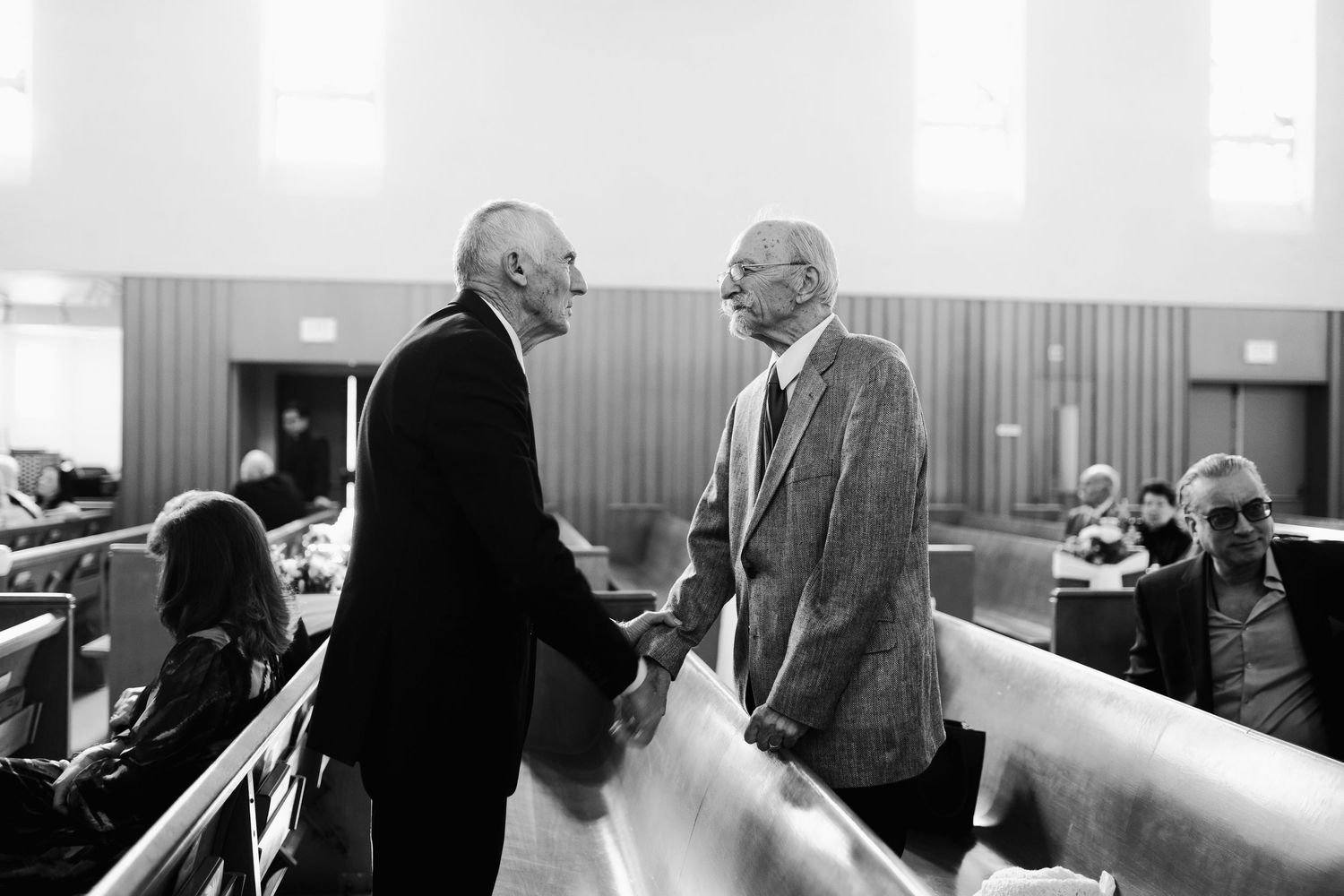 Two elderly men shake hands and converse in a church, captured in black and white photography.