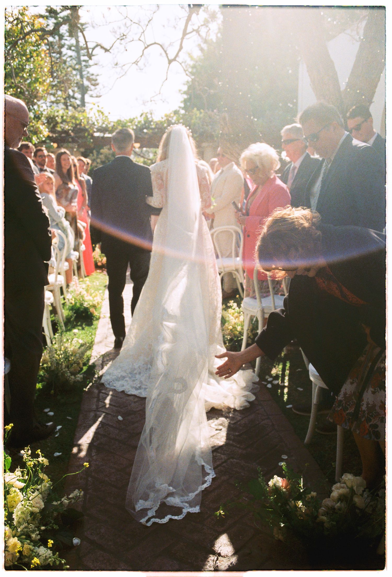 Bride in white gown and long veil walks down outdoor wedding aisle surrounded by seated guests in sunlight.