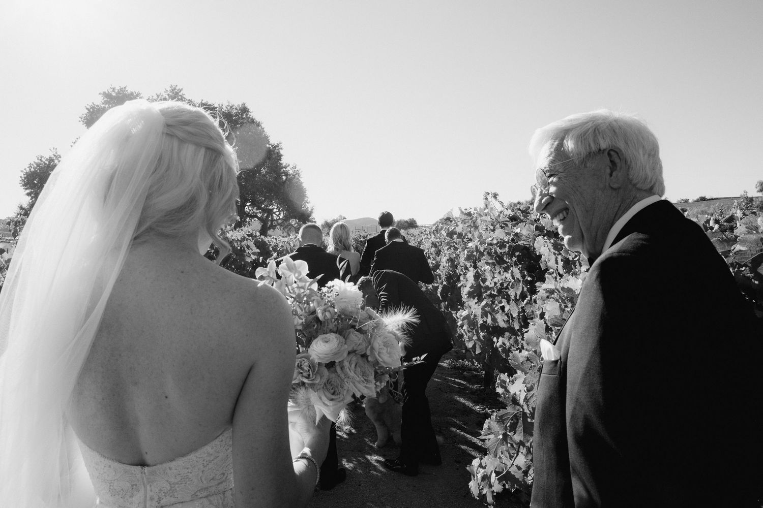 Black and white photo of a bride in veil holding bouquet standing with elderly man at outdoor wedding ceremony.