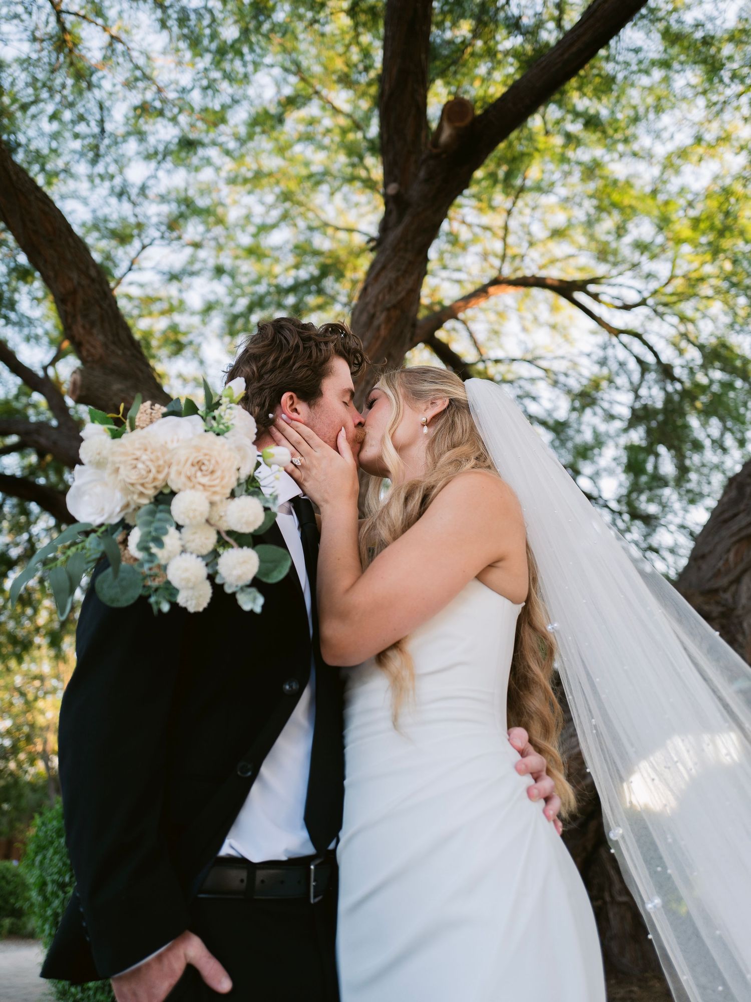 Bride and groom share a passionate kiss under a large tree, bride holding white floral bouquet.