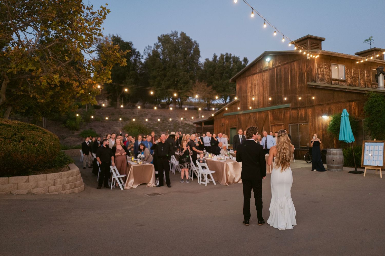 Bride and groom share a first dance on an outdoor patio with guests seated under warm string lights at dusk.