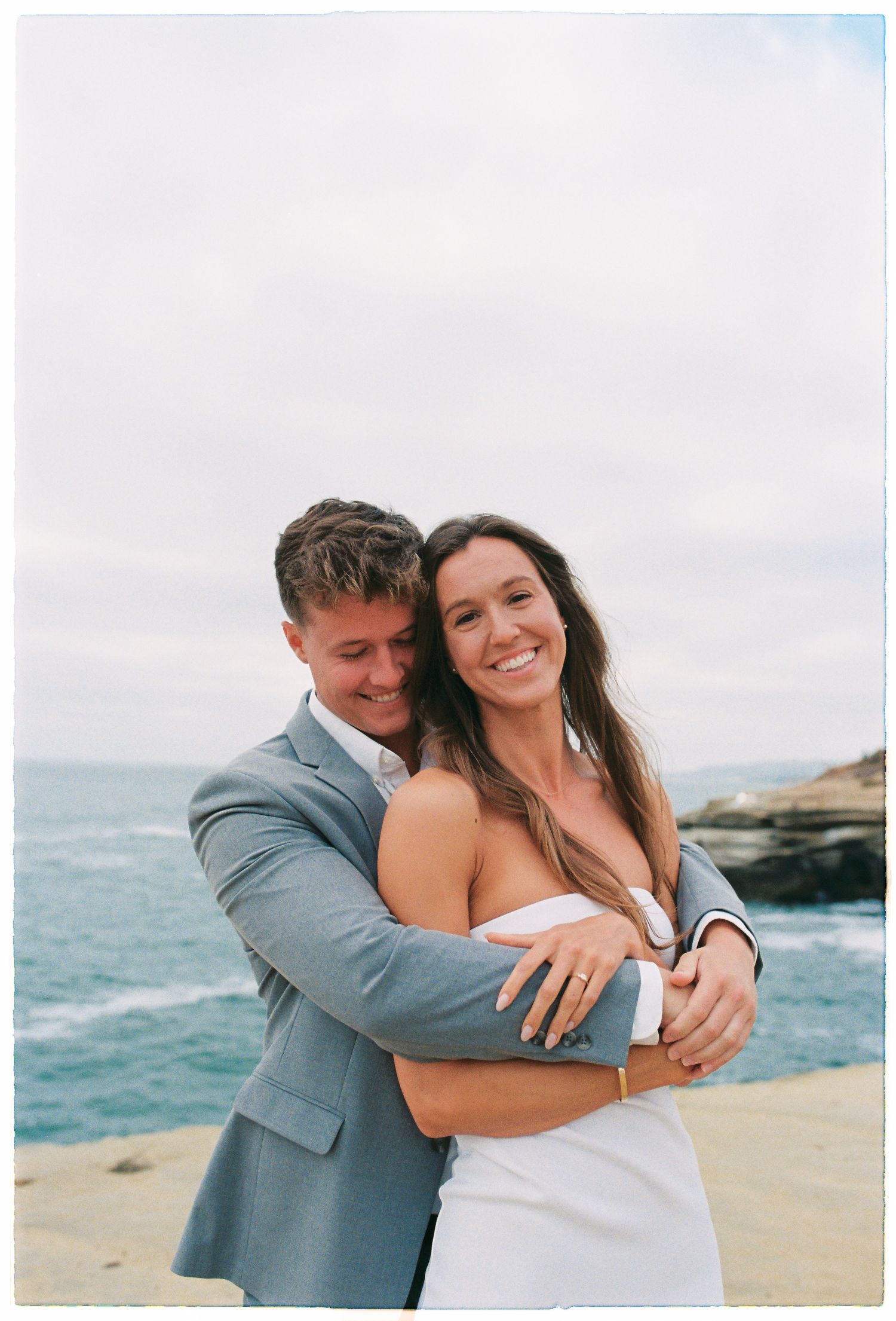 Smiling bride in white strapless dress embraced from behind by groom in gray suit near ocean cliffs on overcast day.