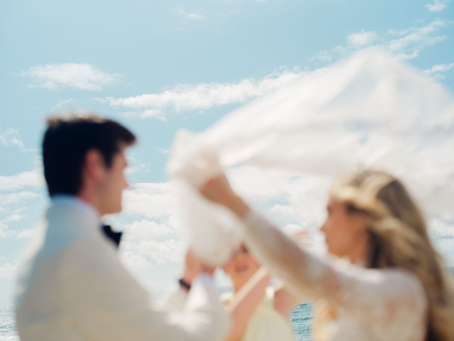 Blurred artistic photo of bride and groom playing with veil against a bright blue sky backdrop.