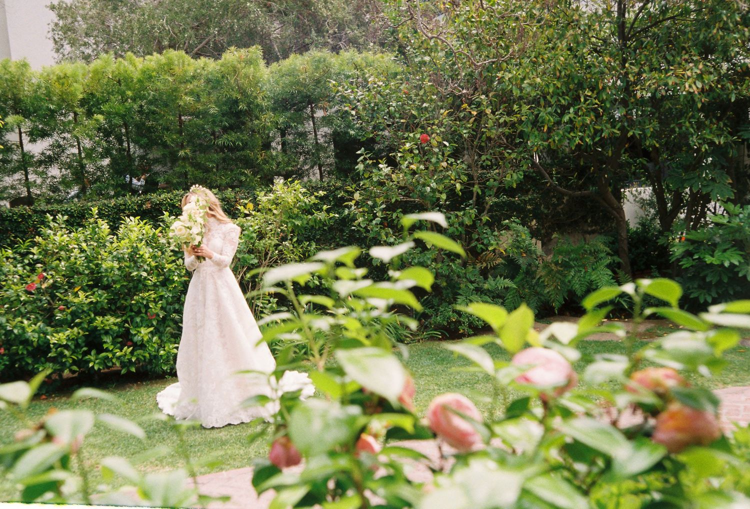 Bride in a long lace wedding gown walks through a lush green garden holding a floral bouquet.