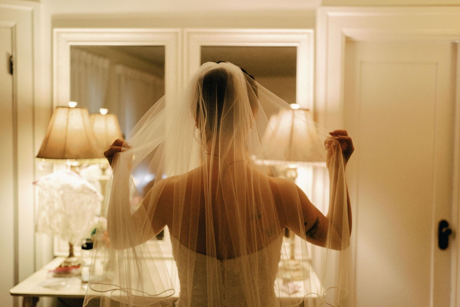 Bride seen from behind holding her wedding veil in a warmly lit dressing room.