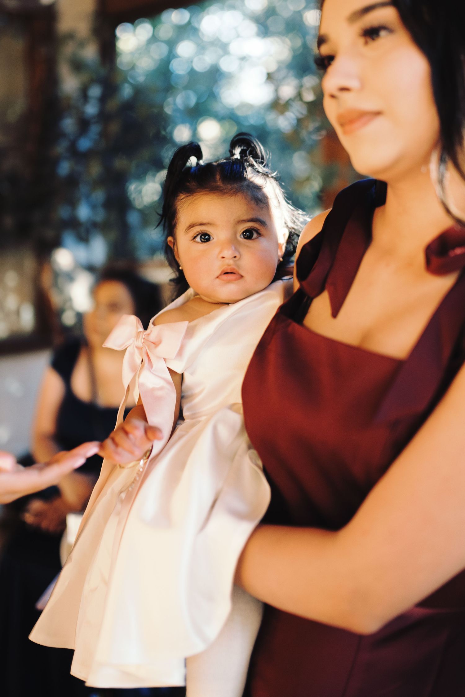 Bridesmaid in burgundy dress holds an adorable baby girl in a white dress with pink bow.