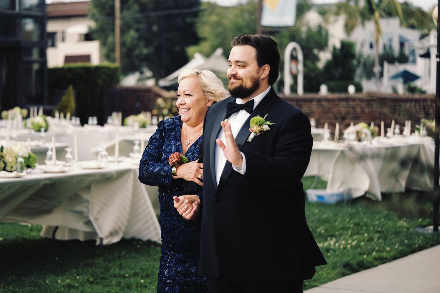 Groom in black tuxedo escorting mother in blue sequin dress at outdoor wedding reception.