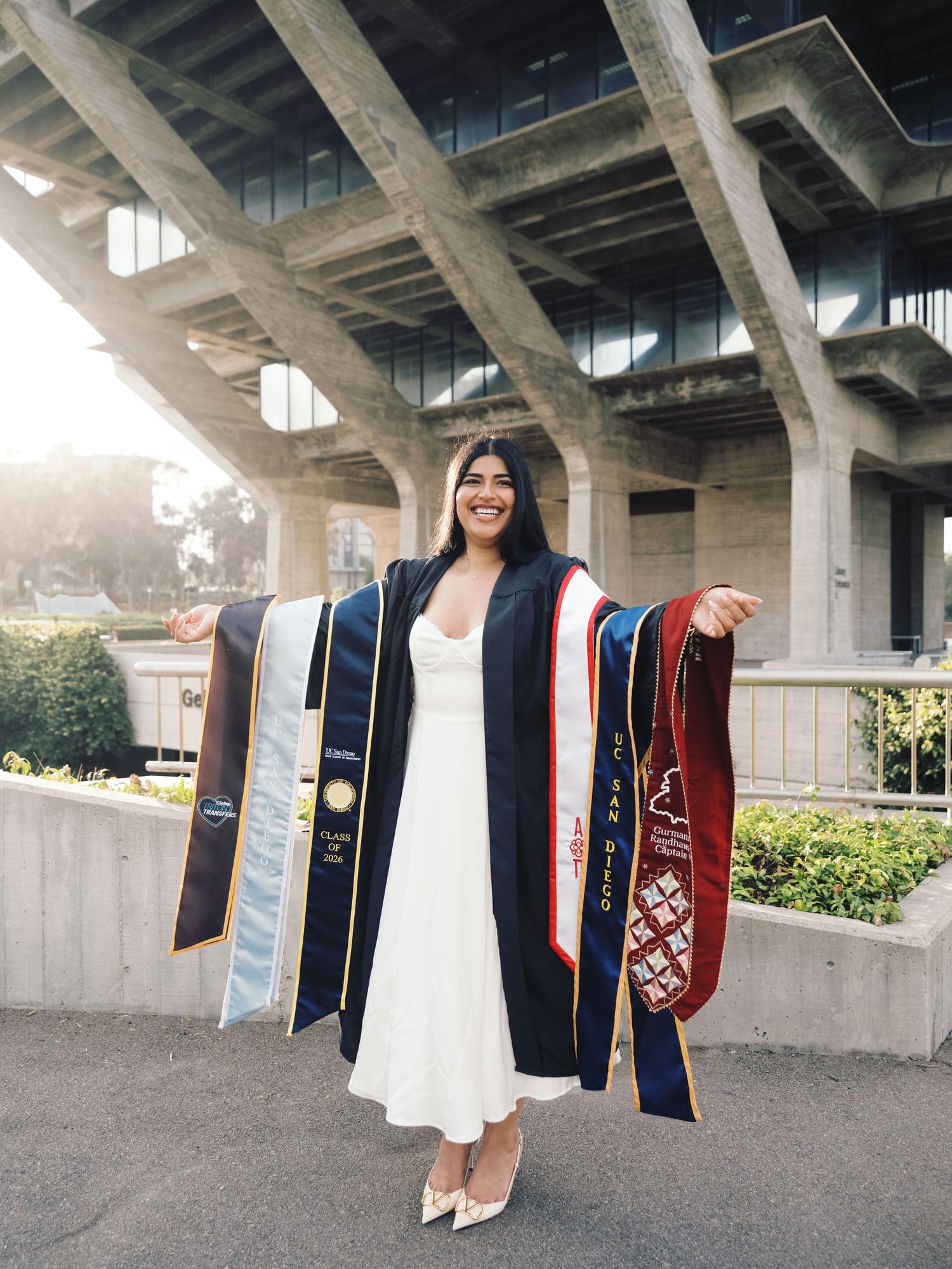 Graduate poses in white dress displaying multiple colorful stoles under modern concrete structure.