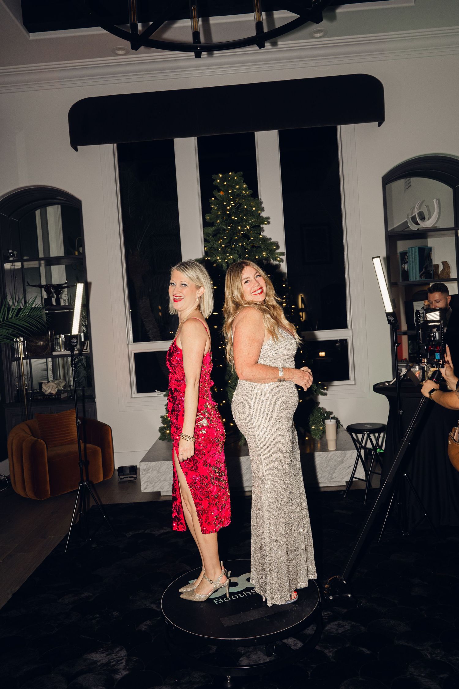 Two women in elegant gowns pose back-to-back at a festive holiday party with a Christmas tree backdrop.