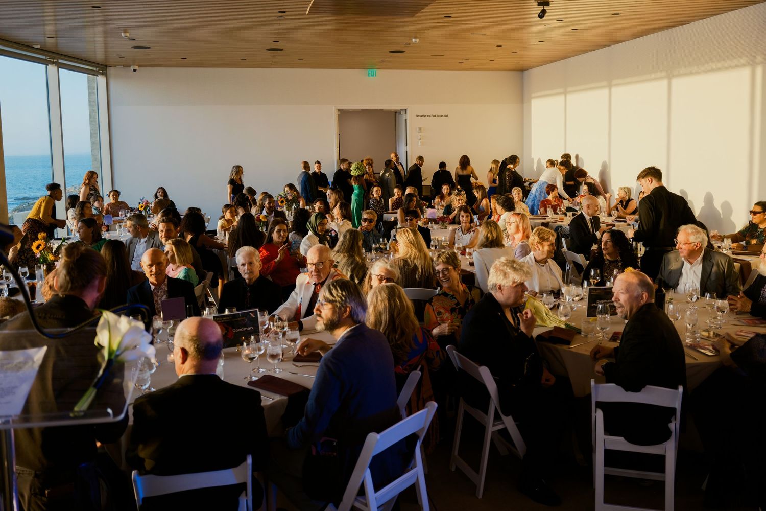 Elegant banquet hall filled with guests dining at round tables, featuring large windows with ocean views at sunset.