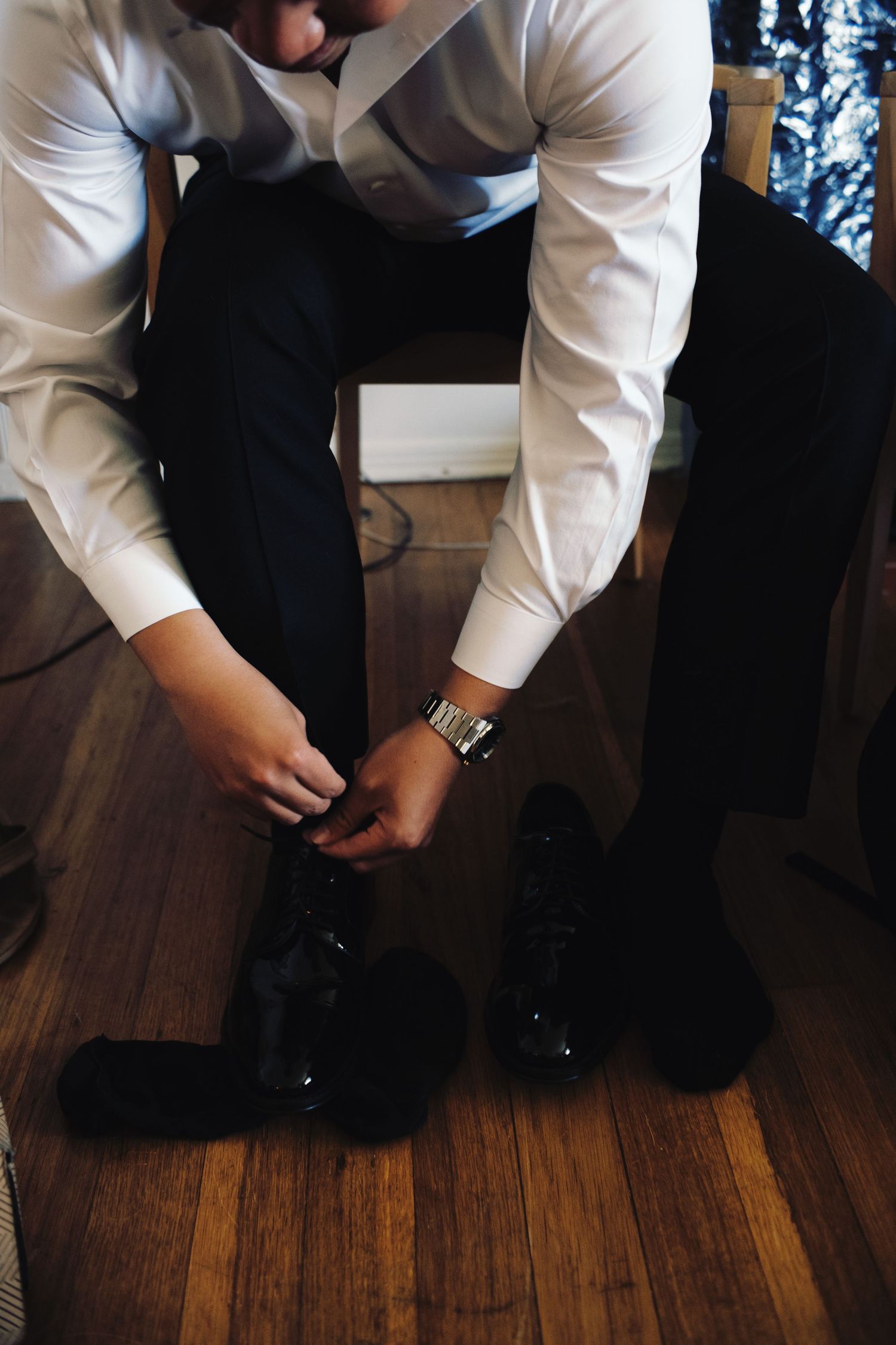 Groom in white dress shirt laces up polished black shoes on hardwood floor before wedding.