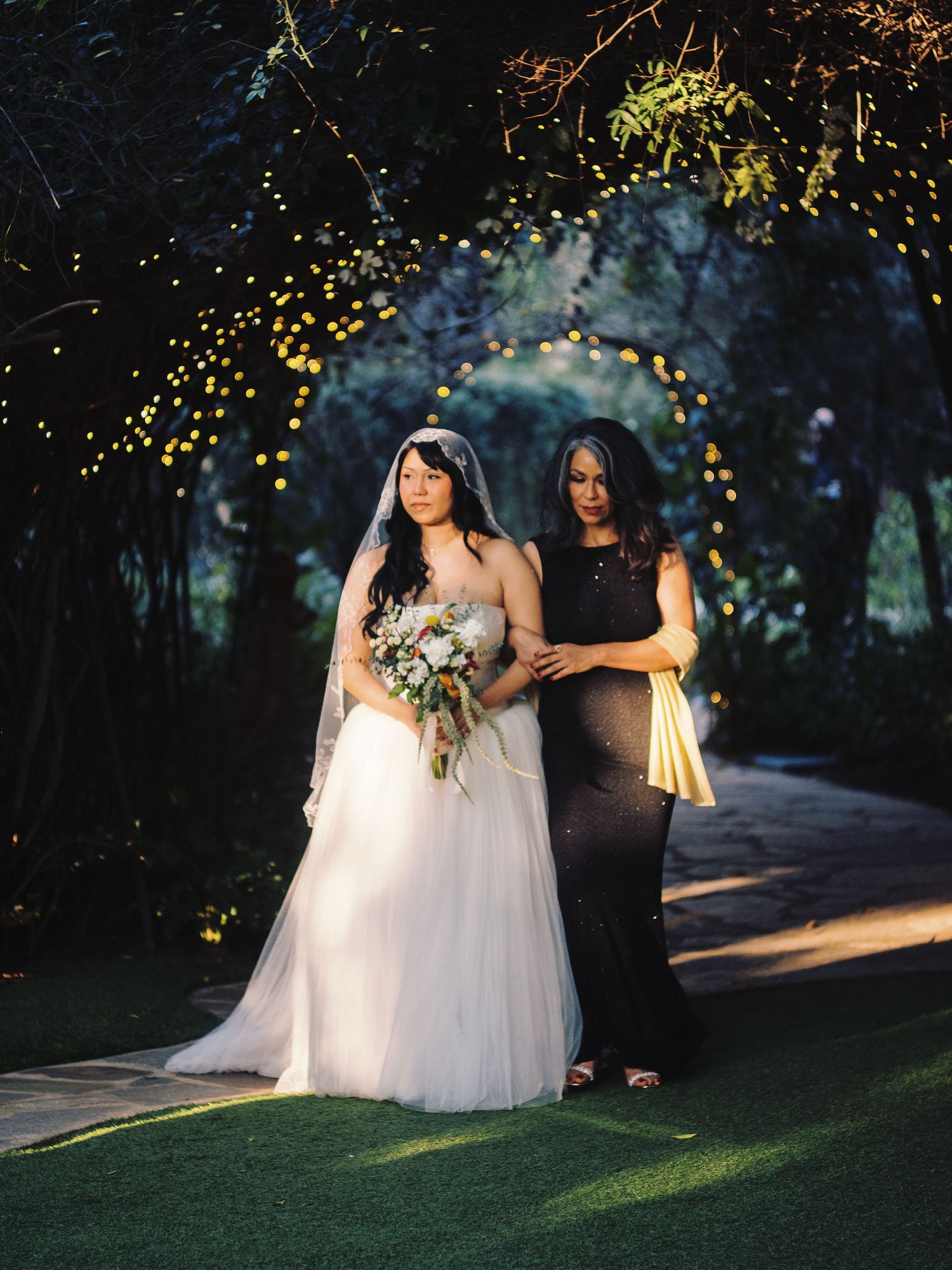 Bride in white gown holding bouquet walks with woman in black dress under fairy lights.