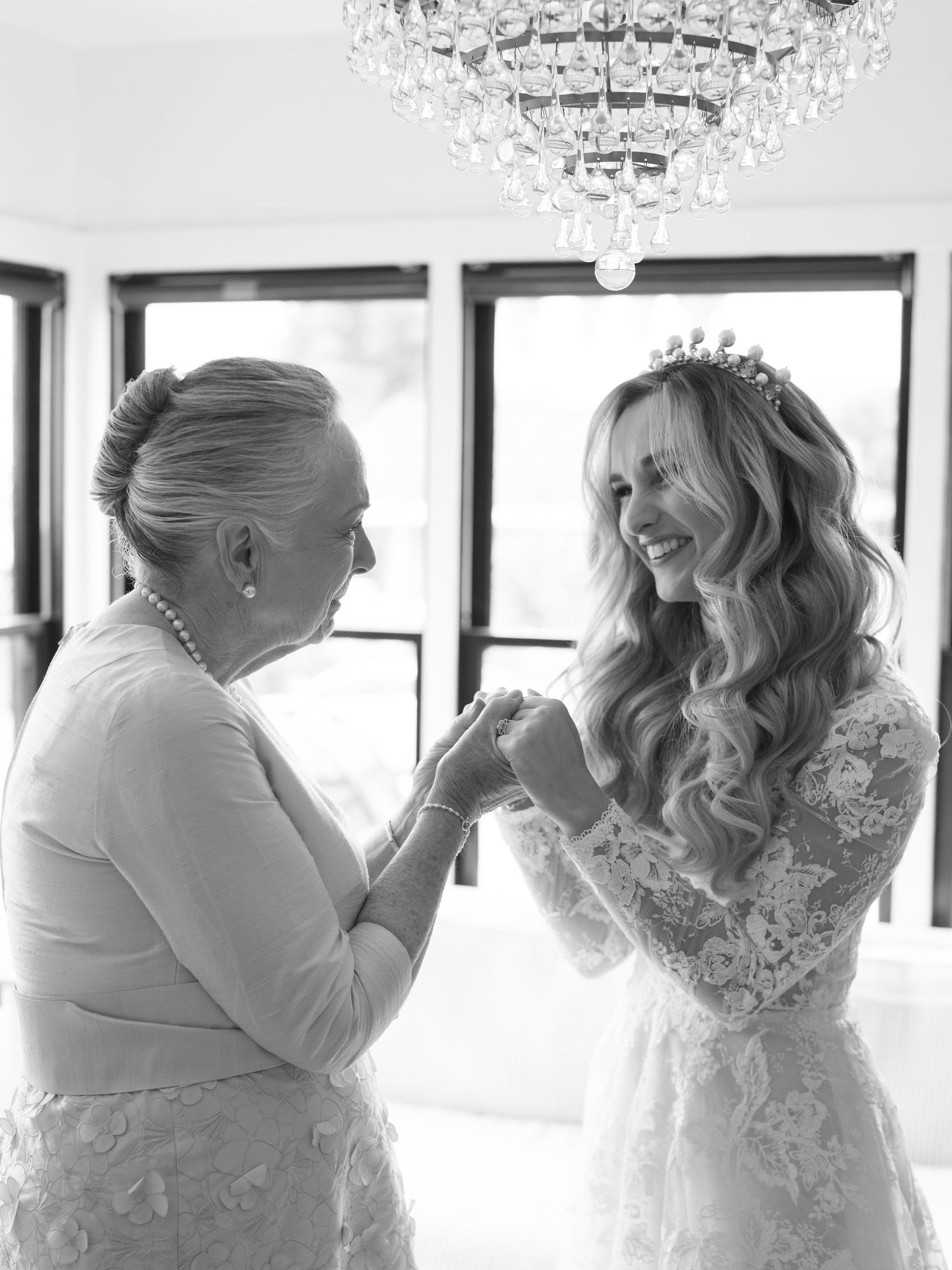 Black and white photo of smiling bride in lace gown and tiara holding hands with older woman under chandelier.