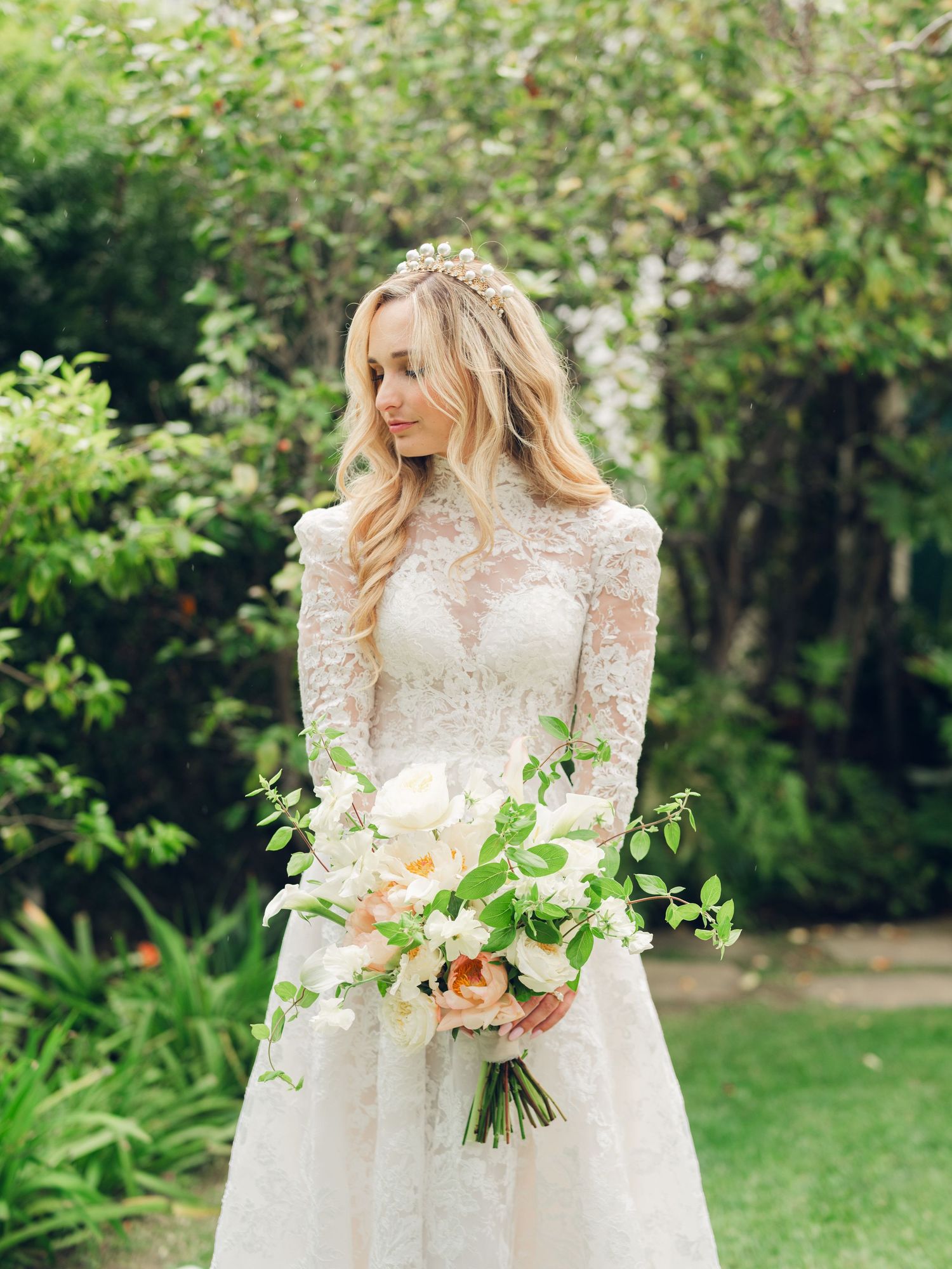 Elegant bride in long-sleeve lace gown holds a bouquet of blush roses and greenery in a garden.
