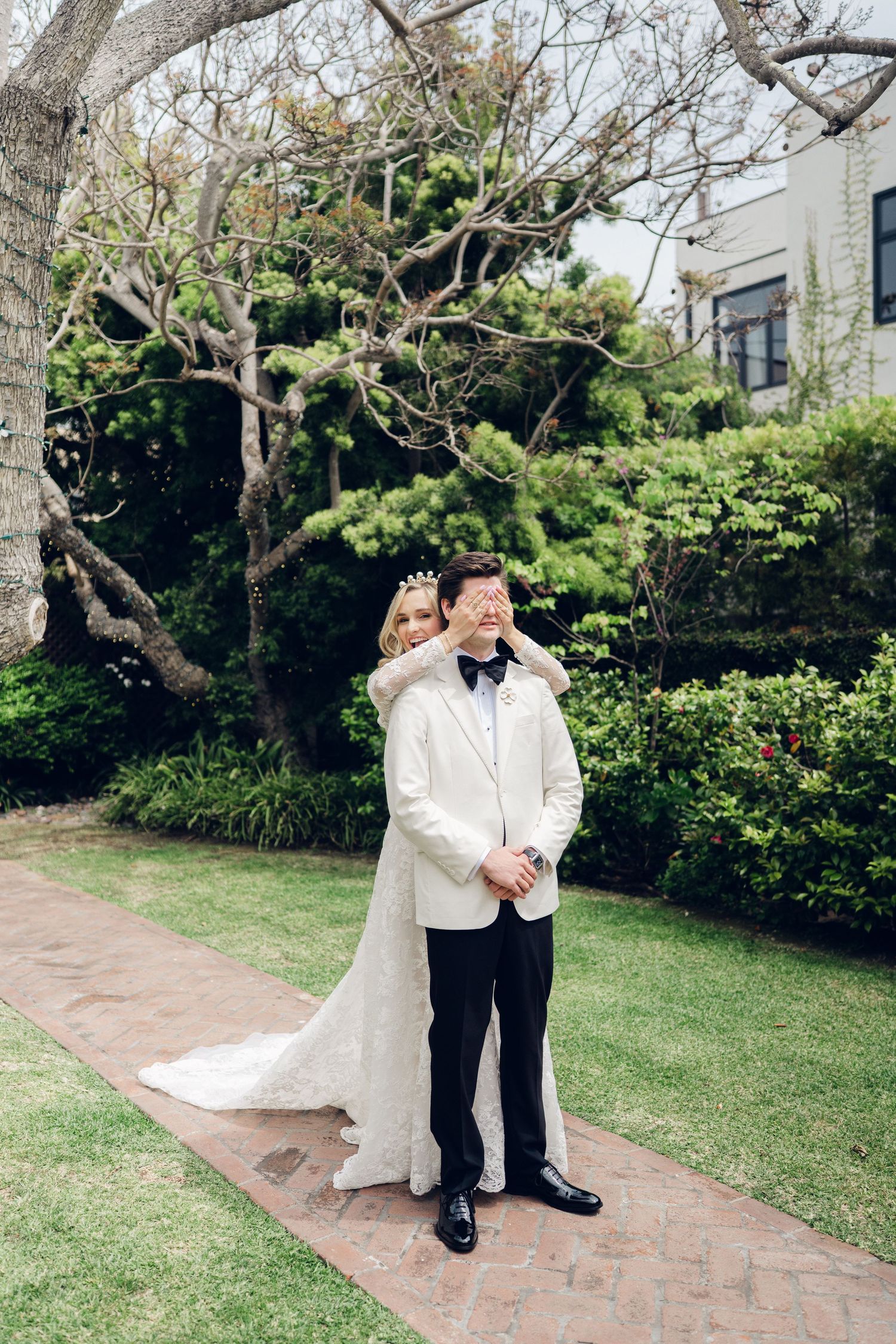 Bride surprises groom by covering his eyes during romantic first look moment in garden setting.