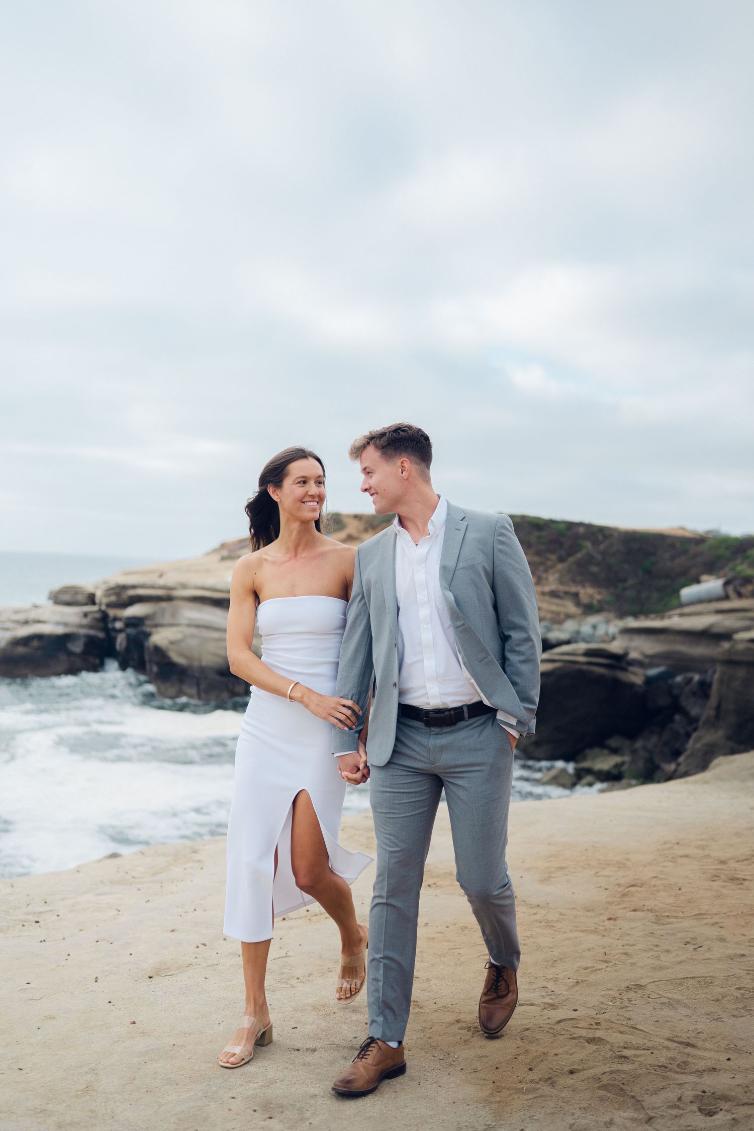 Couple walking on sandy beach, woman in white strapless dress and man in gray suit, smiling at each other with rocky cliffs behind.