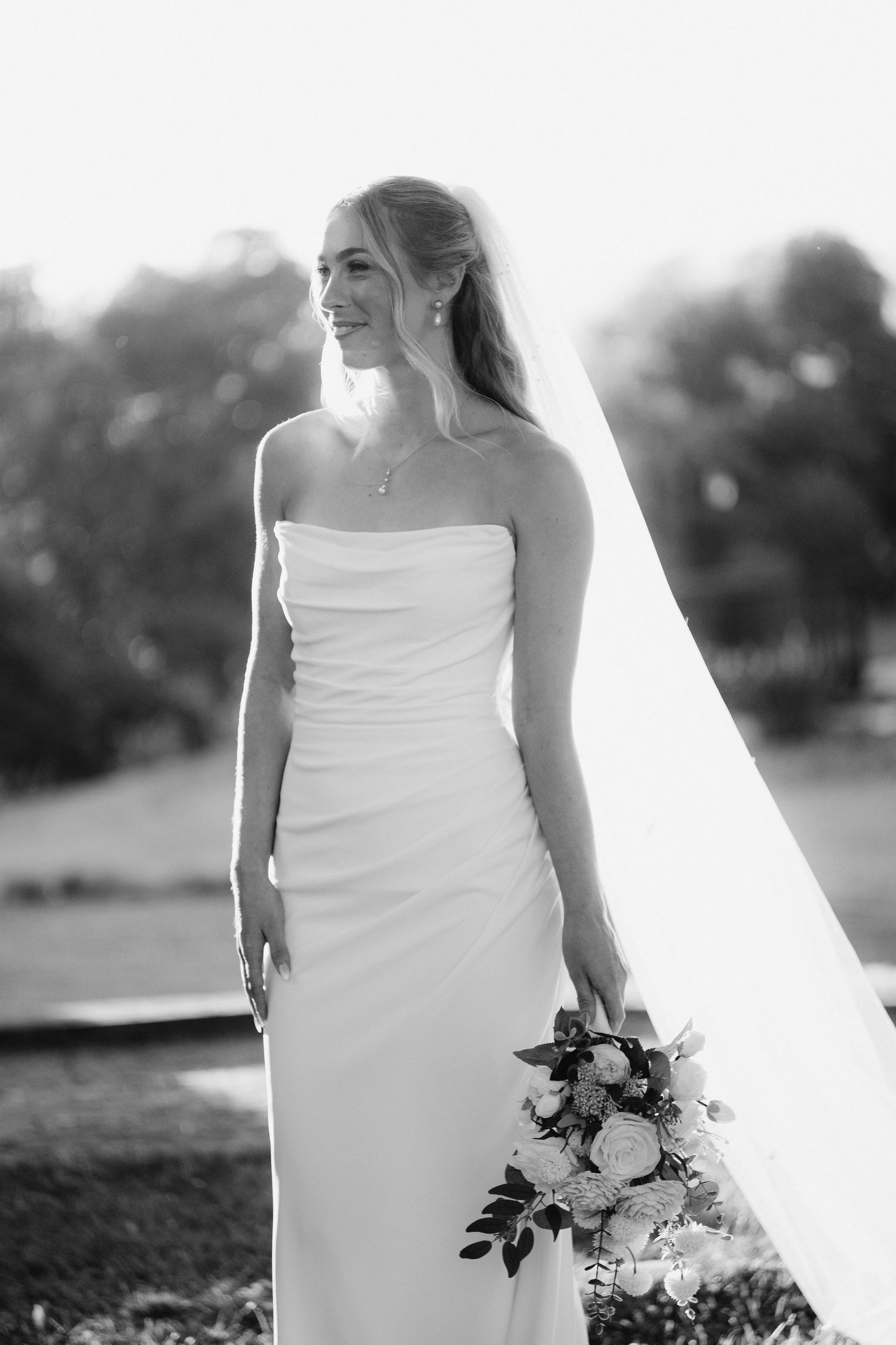 Elegant bride in strapless white gown holds a bouquet while her veil flows in a black and white portrait.