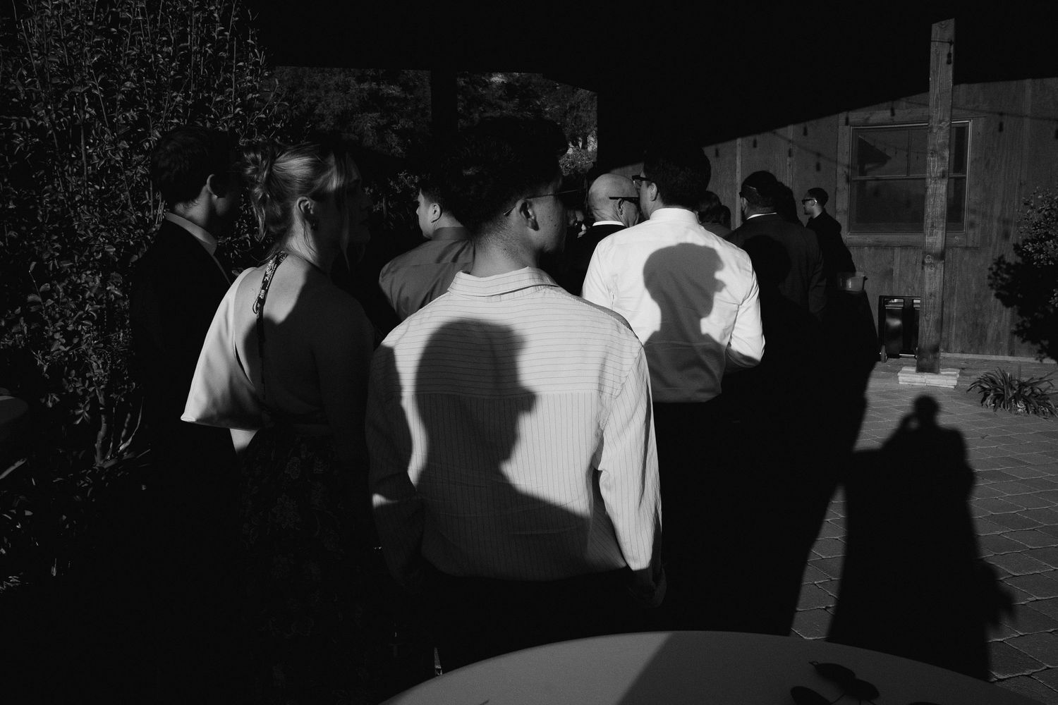 Black and white photo of wedding guests gathered outdoors at night with dramatic shadows cast on a wall.